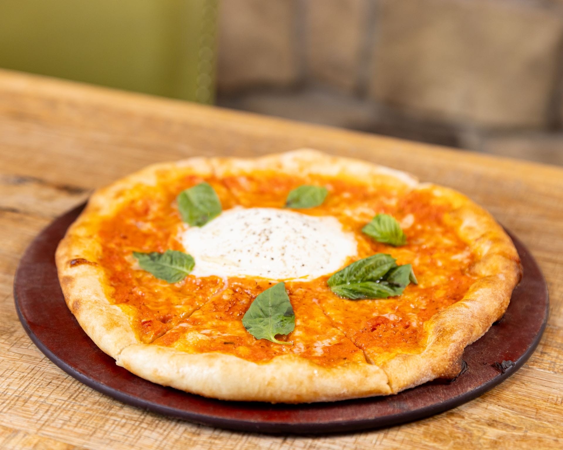 A pizza with melted cheese, tomato sauce, fresh basil leaves, and a dollop of cheese in the center on a wooden table.