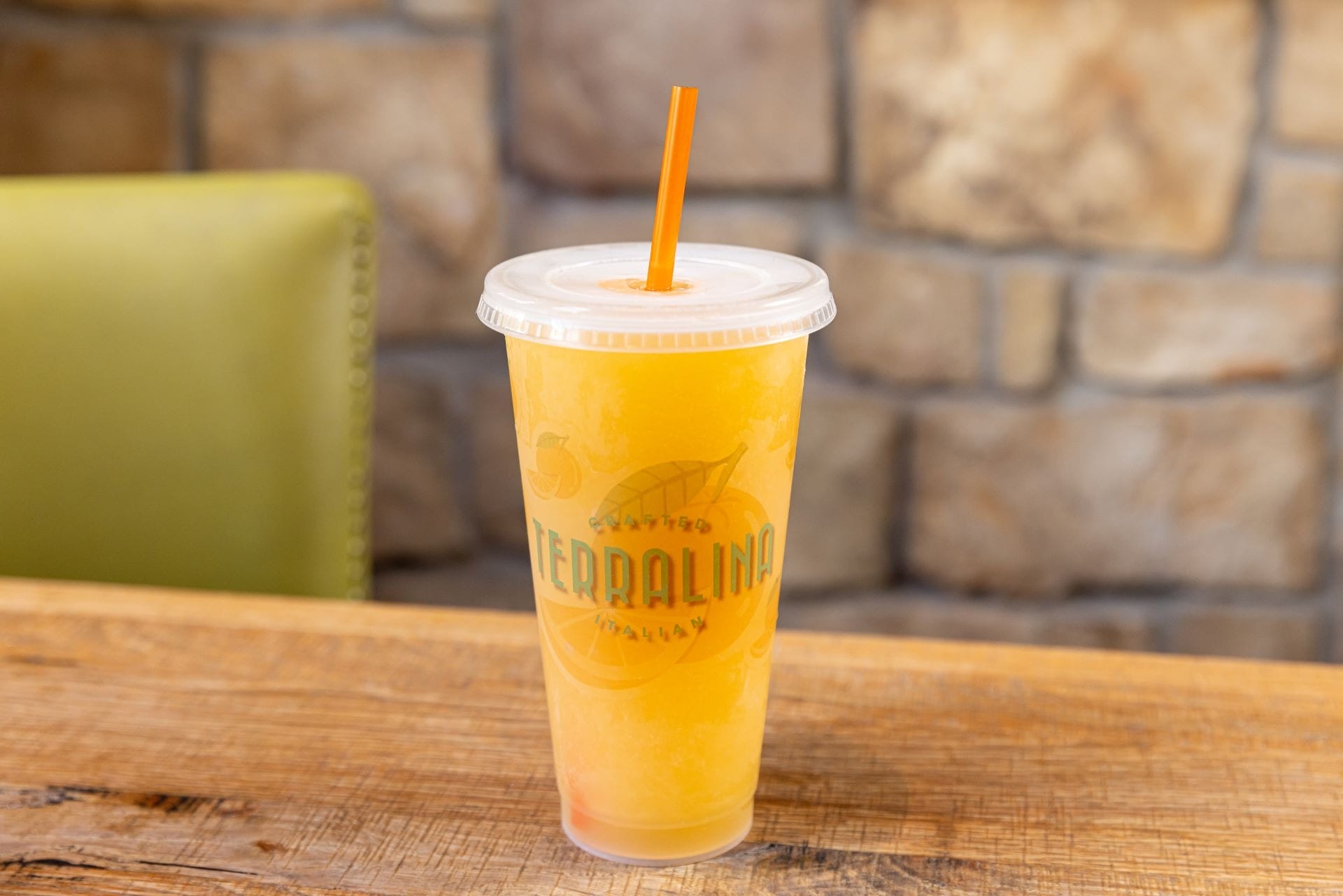 A plastic cup of orange slush with an orange straw sits on a wooden table in front of a stone wall.