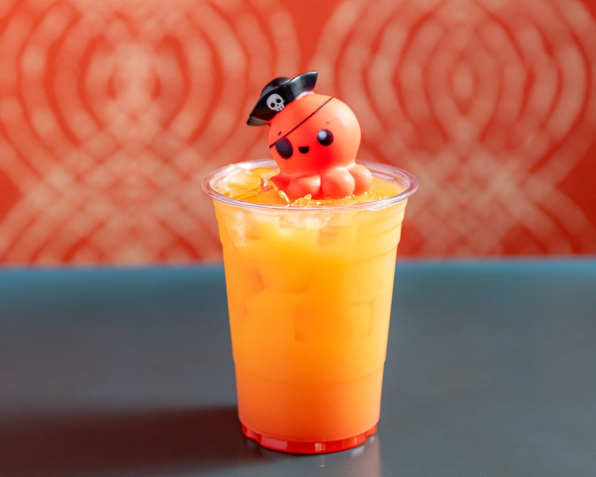 An orange drink with ice topped with a cute pirate octopus decoration in a clear plastic cup.