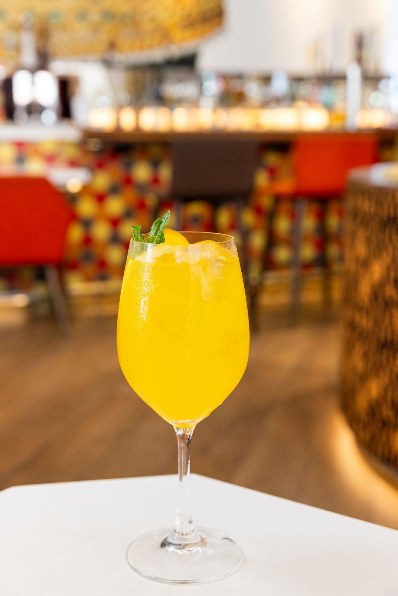 A yellow cocktail with ice and mint garnish in a wine glass on a table, with a colorful bar background.