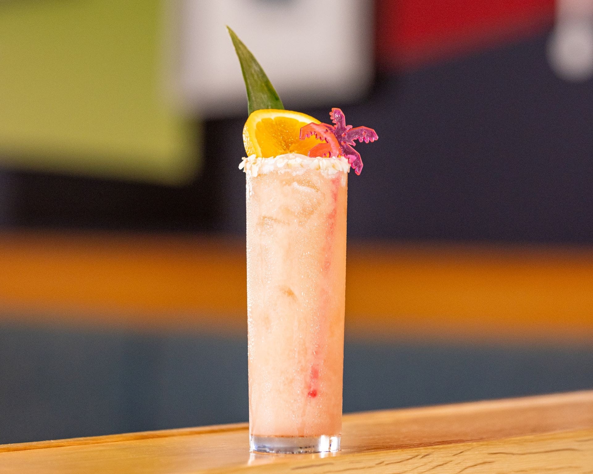 A frosty pink cocktail in a tall glass, garnished with an orange slice, pineapple leaf, whipped cream, and a purple flower.