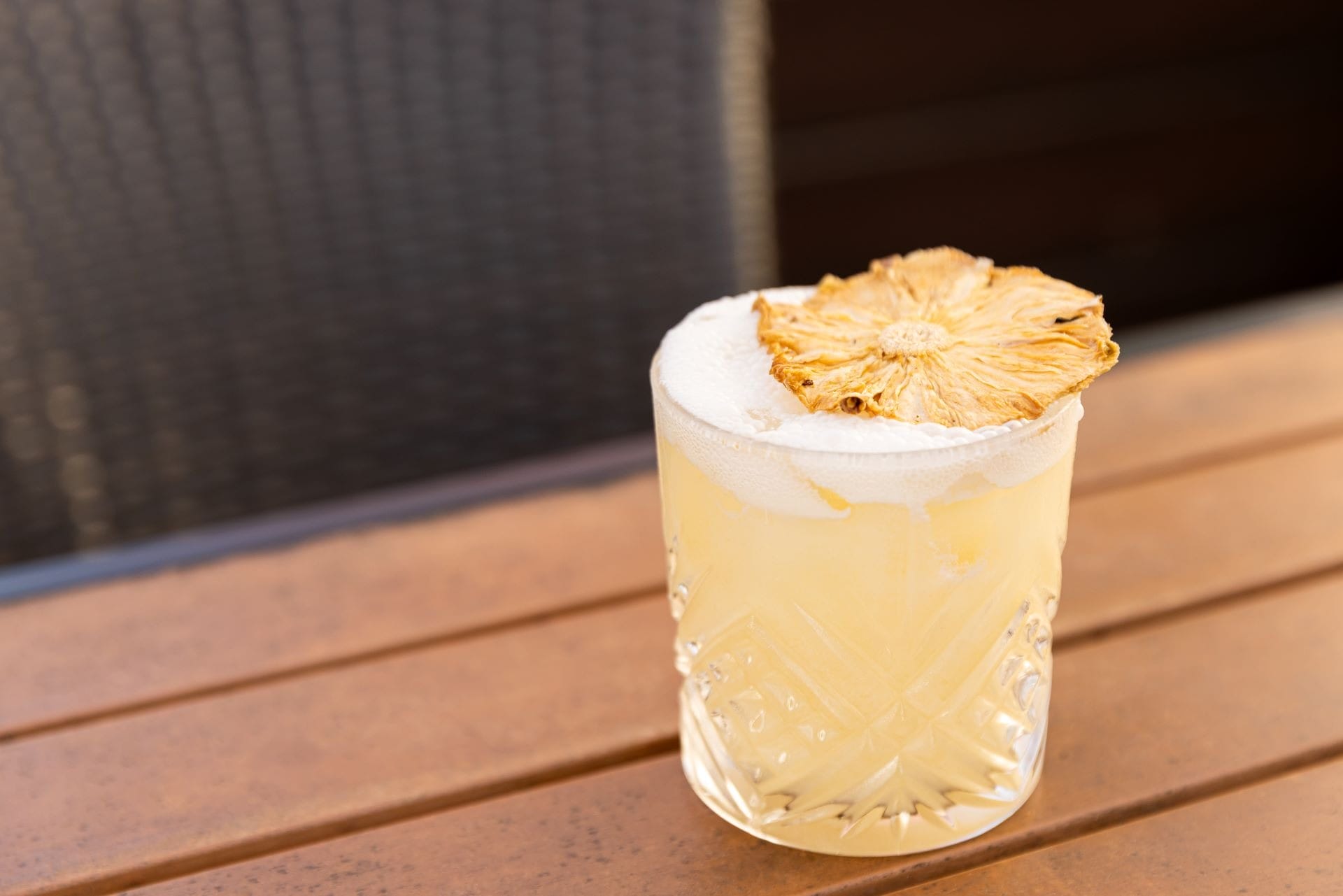 A yellow cocktail in a textured glass, topped with foam and a dried pineapple slice, on a wooden table.
