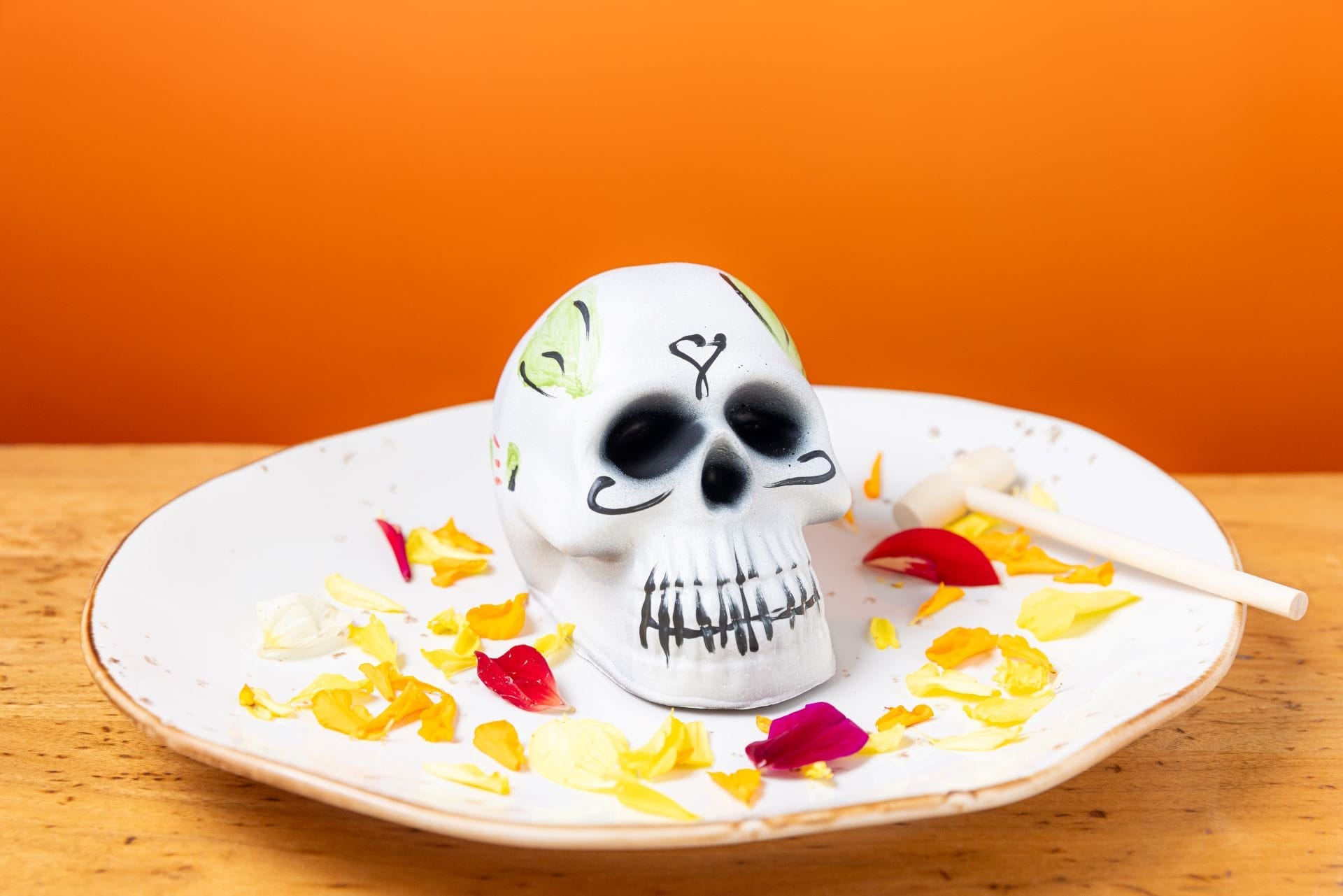 Sugar skull on a plate with flower petals and a small stick, set against an orange background.