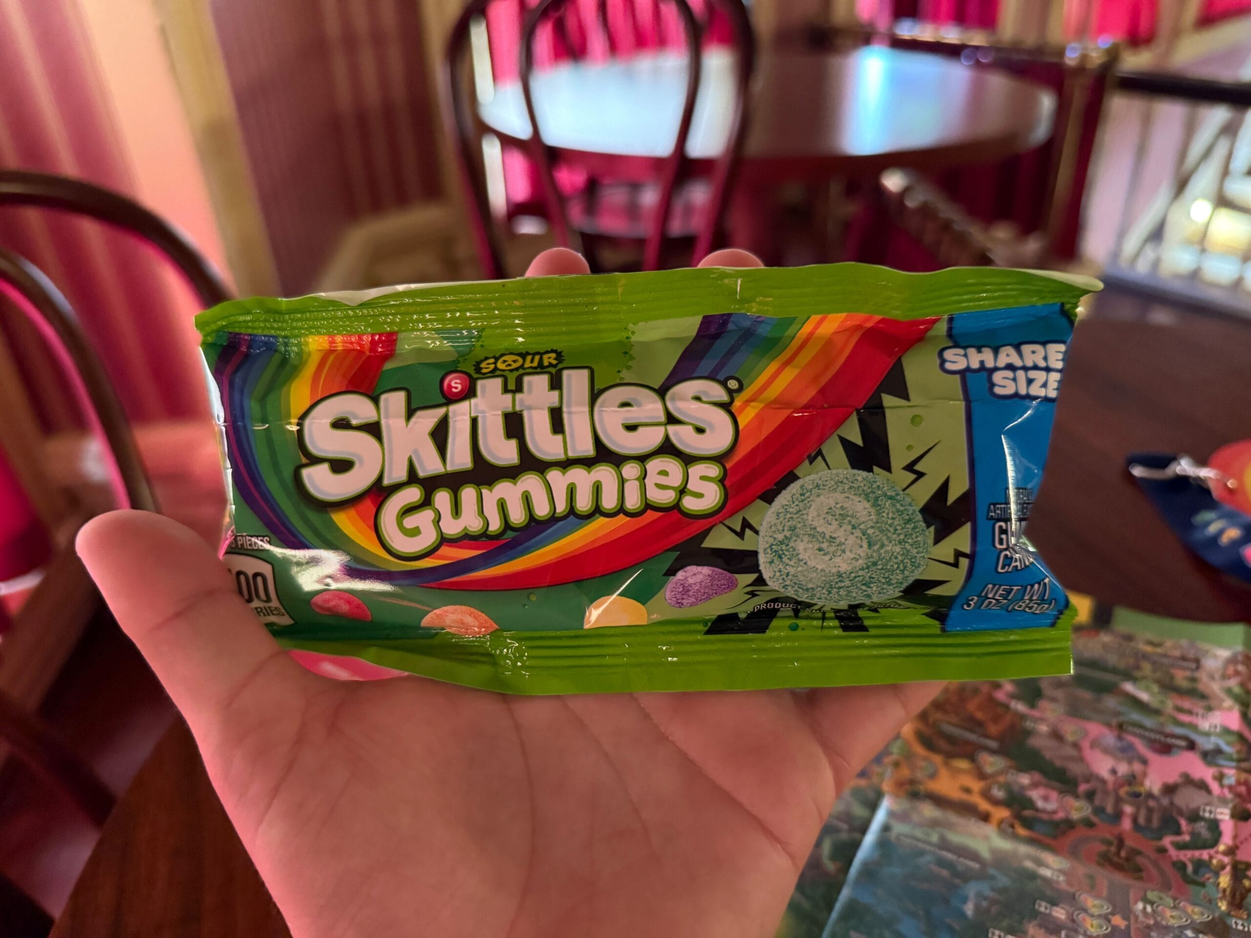 A hand holds Skittles Gummies Sour candy in a colorful room with chairs, tables, and a Disneyland map for 2025 nearby.