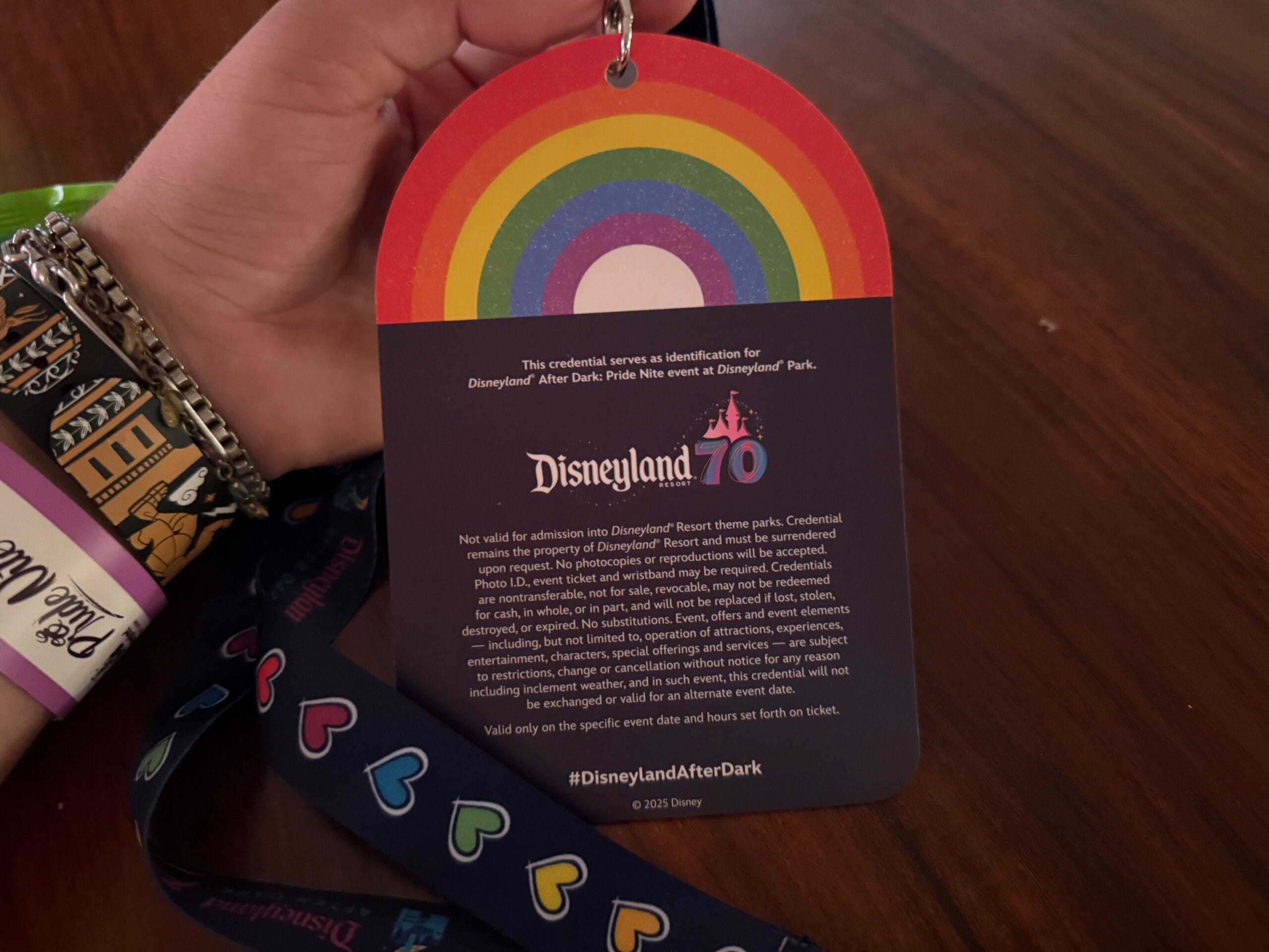 Hand holding a Disneyland Pride Nite lanyard badge with rainbow design, event info, and a glimpse of the Disneyland map.