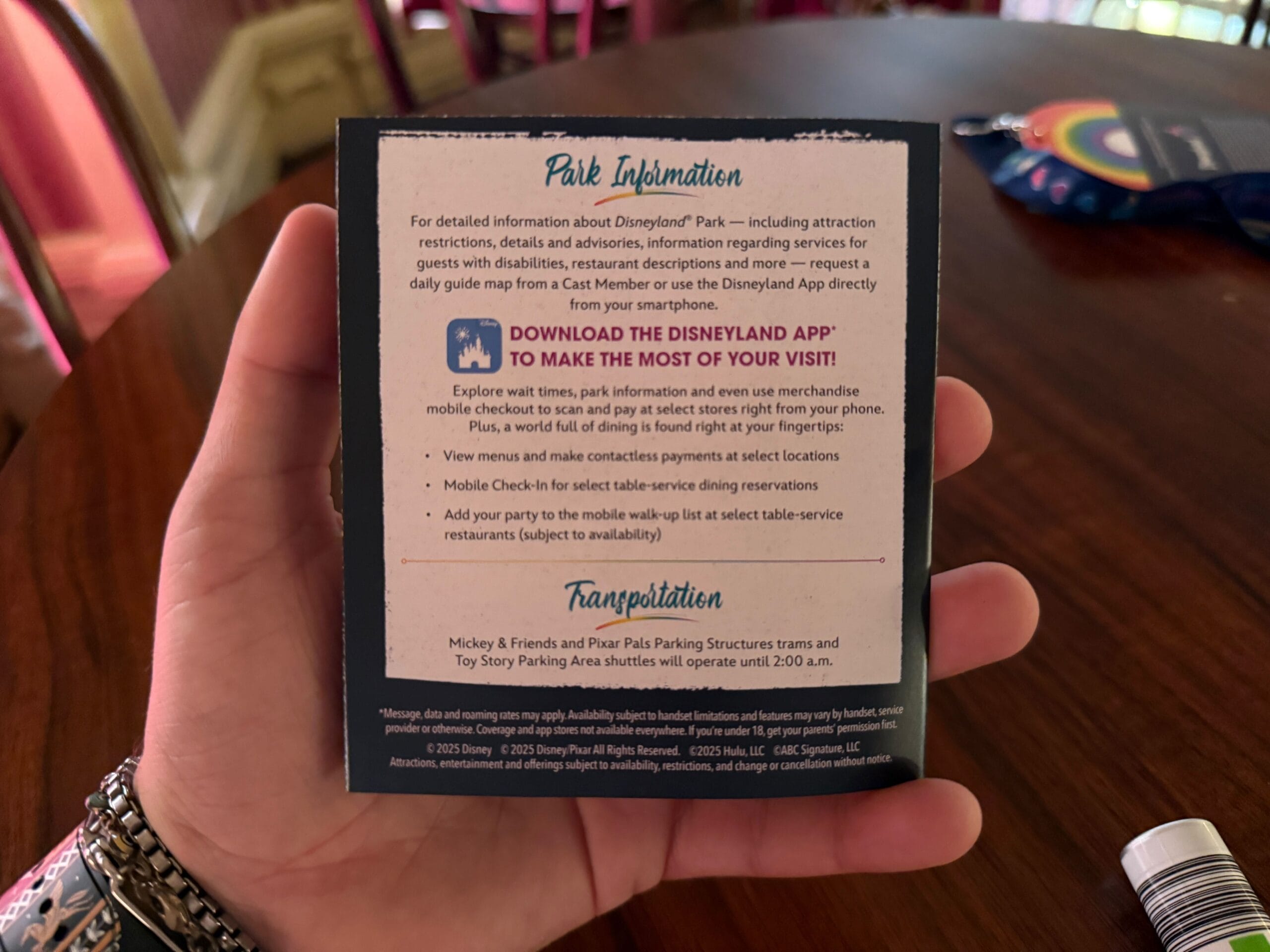 A hand holds a Disneyland Pride Nite 2025 map with park details, app use, and transportation info, indoors.
