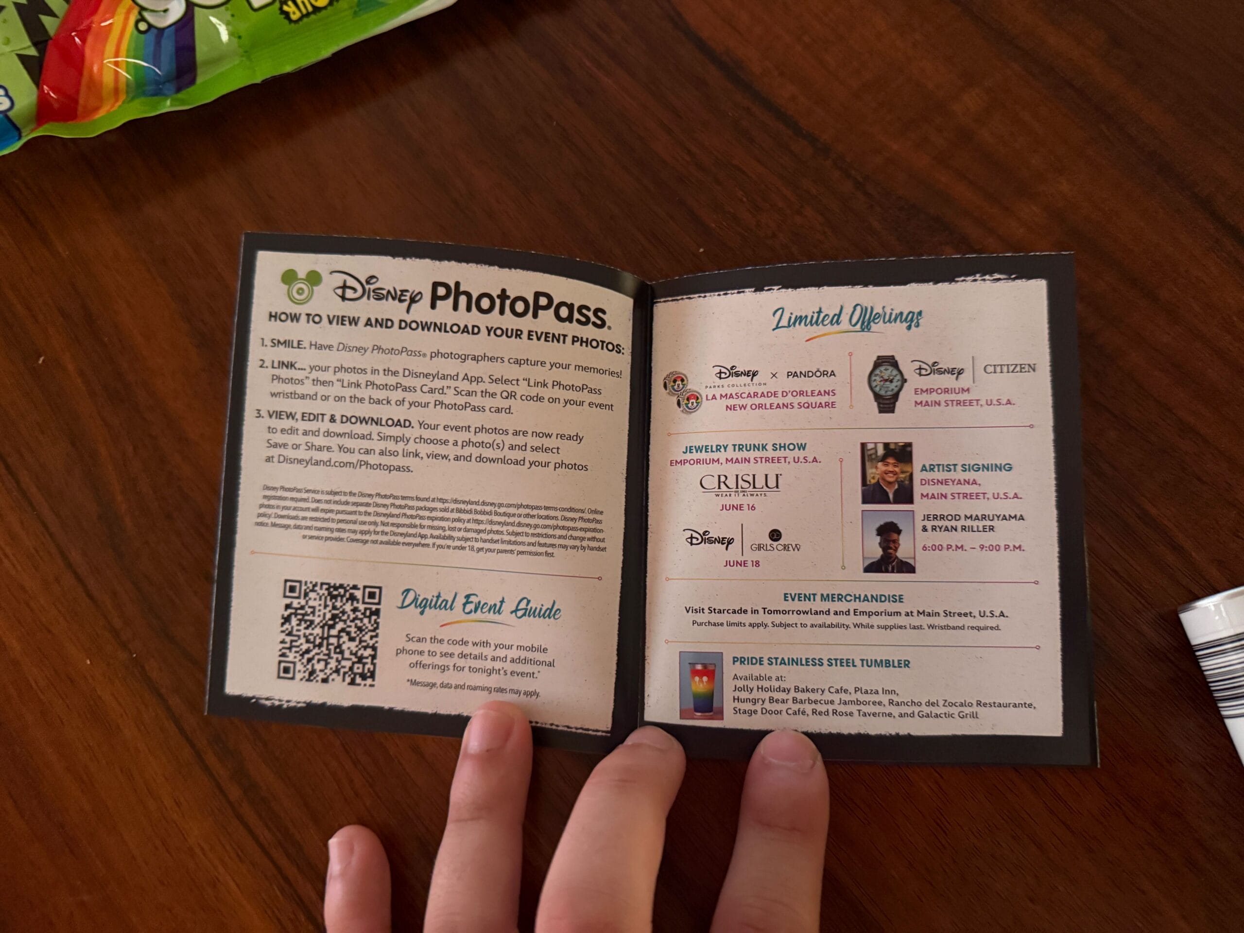 A hand holds open a Disney PhotoPass booklet with info, QR code, and a map at Disneyland Pride Nite on a wooden table.