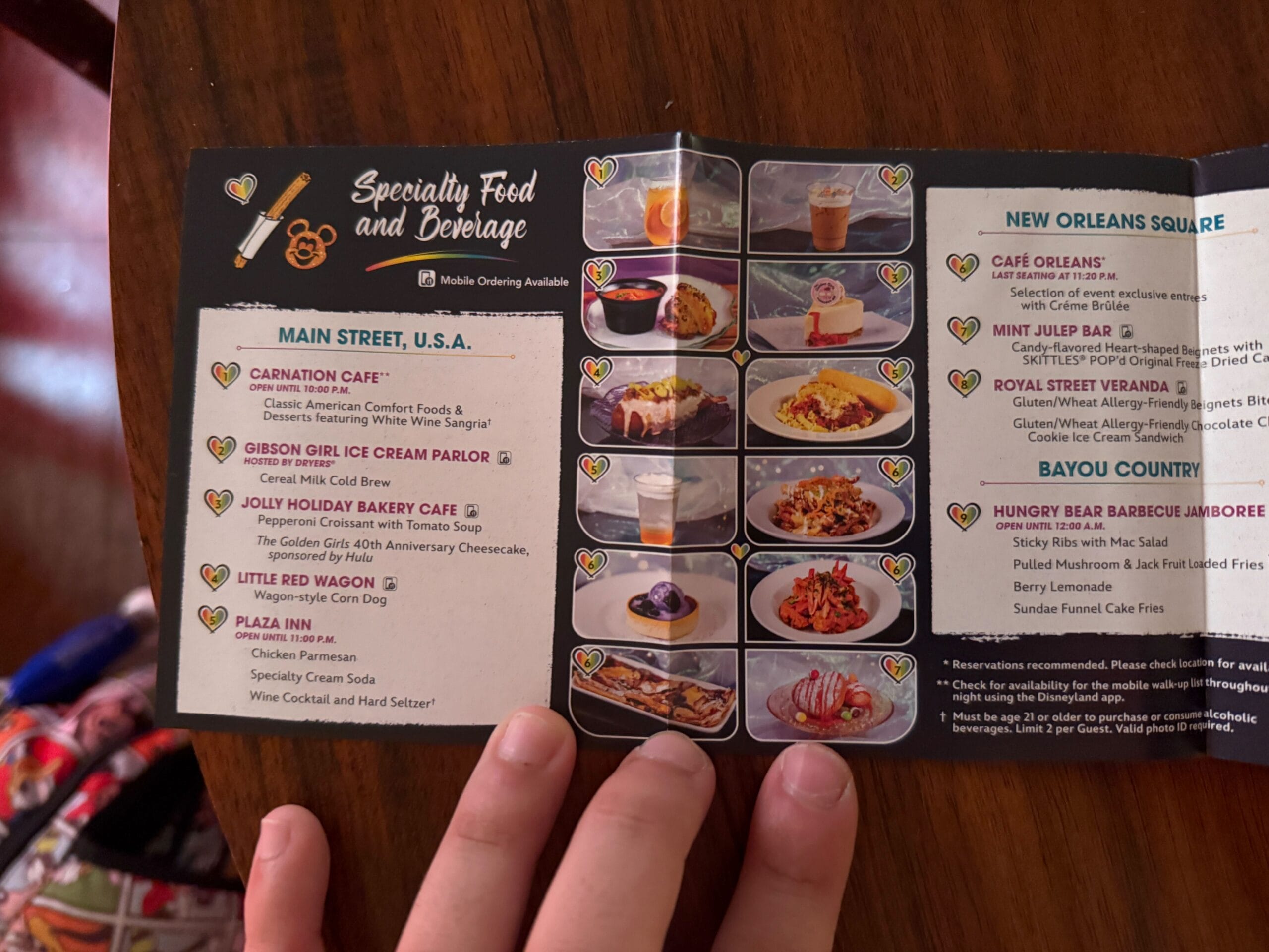 A hand holds a Disneyland Pride Nite menu open, showing food and drink options with images and descriptions.