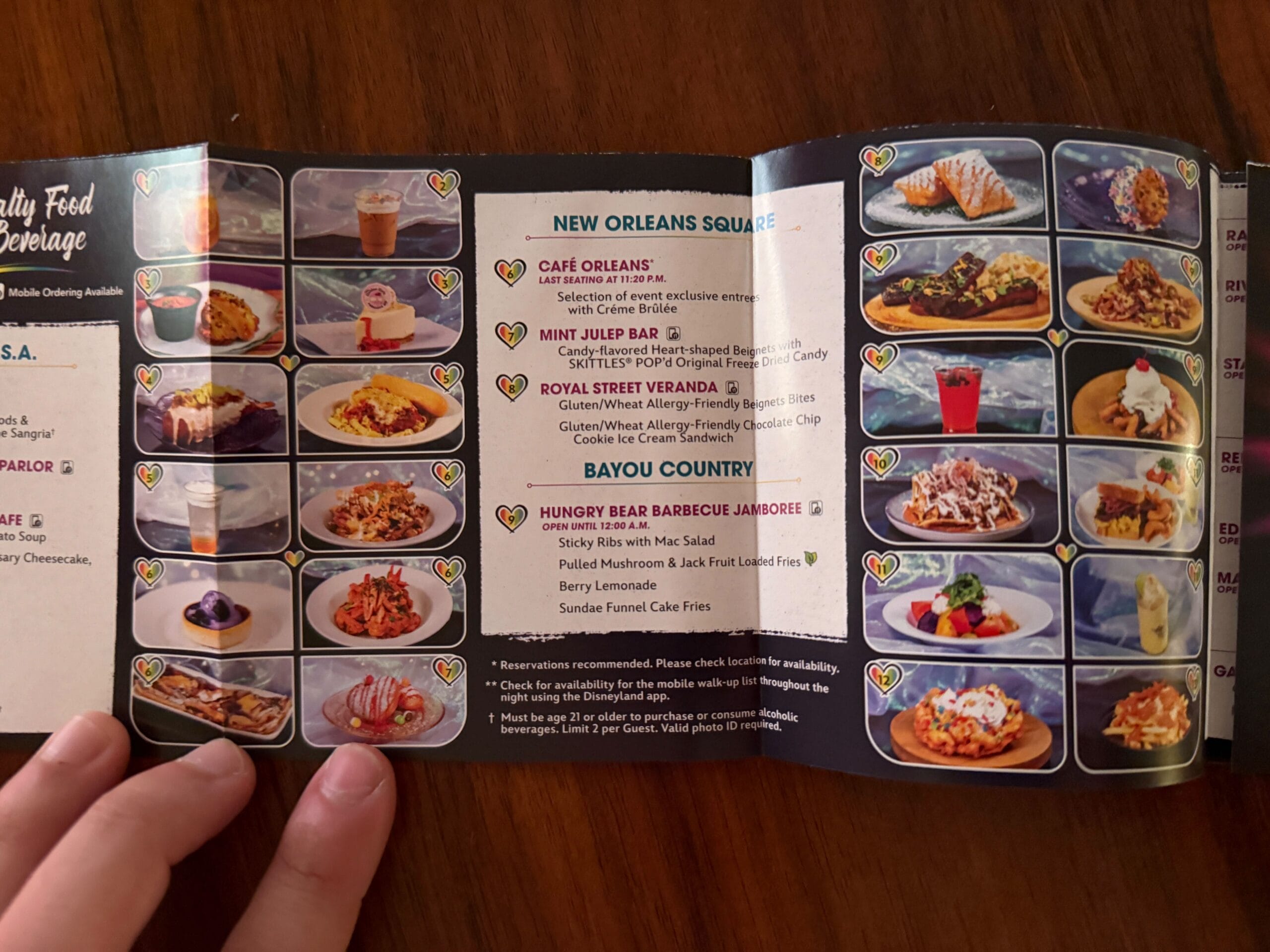 A hand holds a menu and a 2025 map with food and drink photos, spread out on a wooden table.