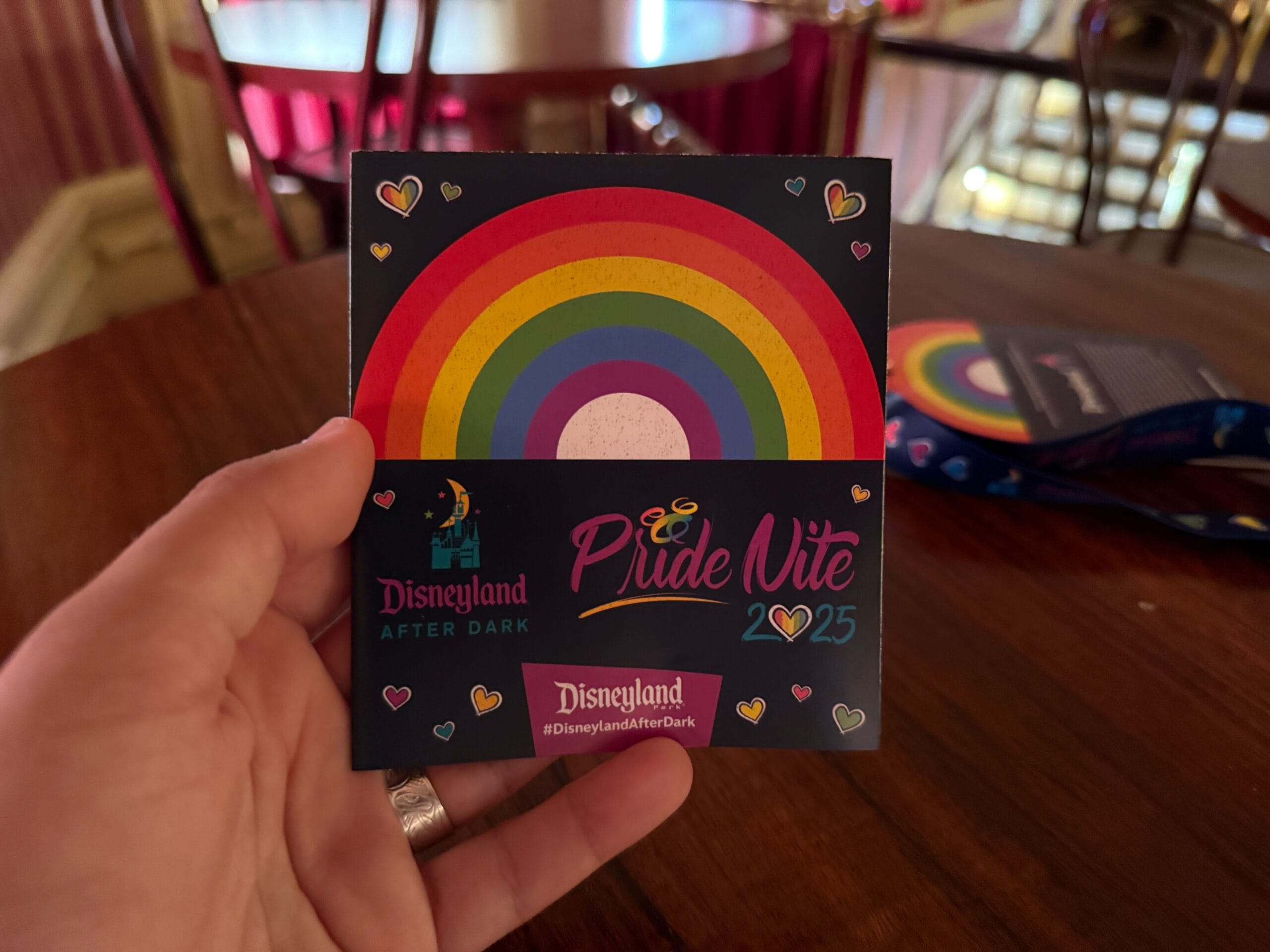 Hand holding a Disneyland Pride Nite 2025 credential with rainbow design, on a table with chairs in the background.