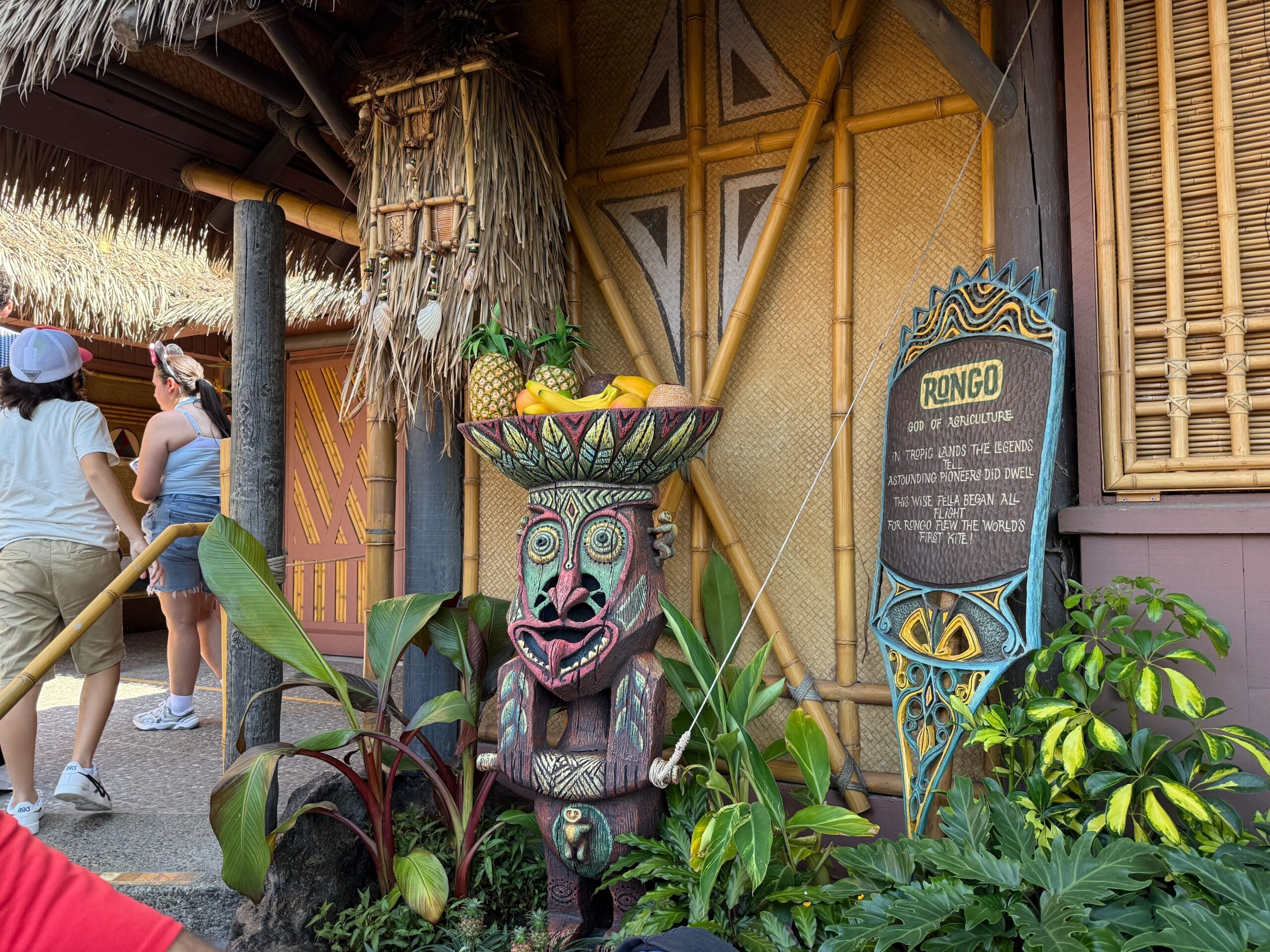 A colorful tiki statue with a fruit bowl stands by a sign in the Enchanted Tiki Garden at Walt Disney.