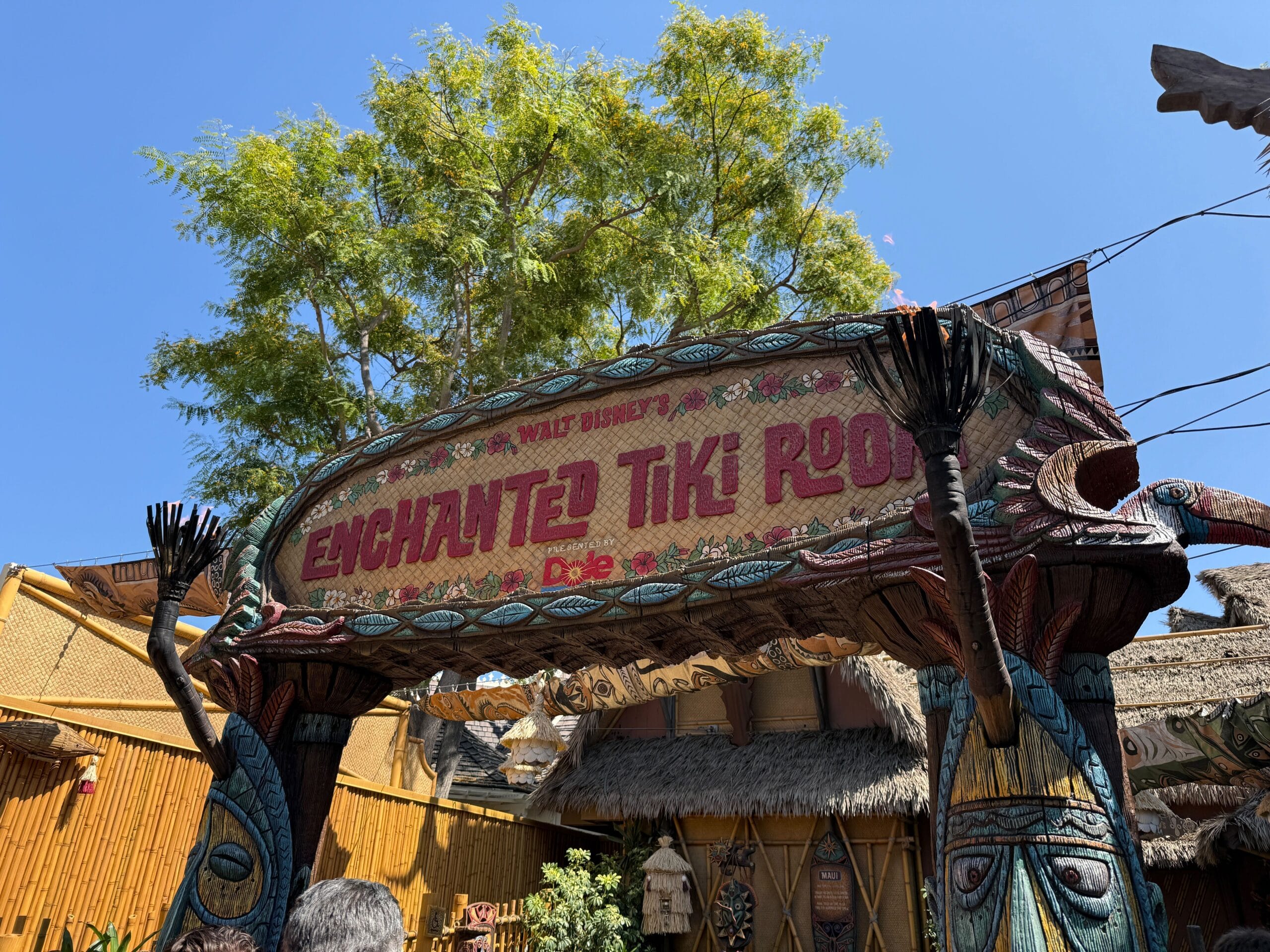 Entrance sign for Walt Disney’s Enchanted Tiki Room in the Pre-Show Area, adorned with tropical carvings under a blue sky.