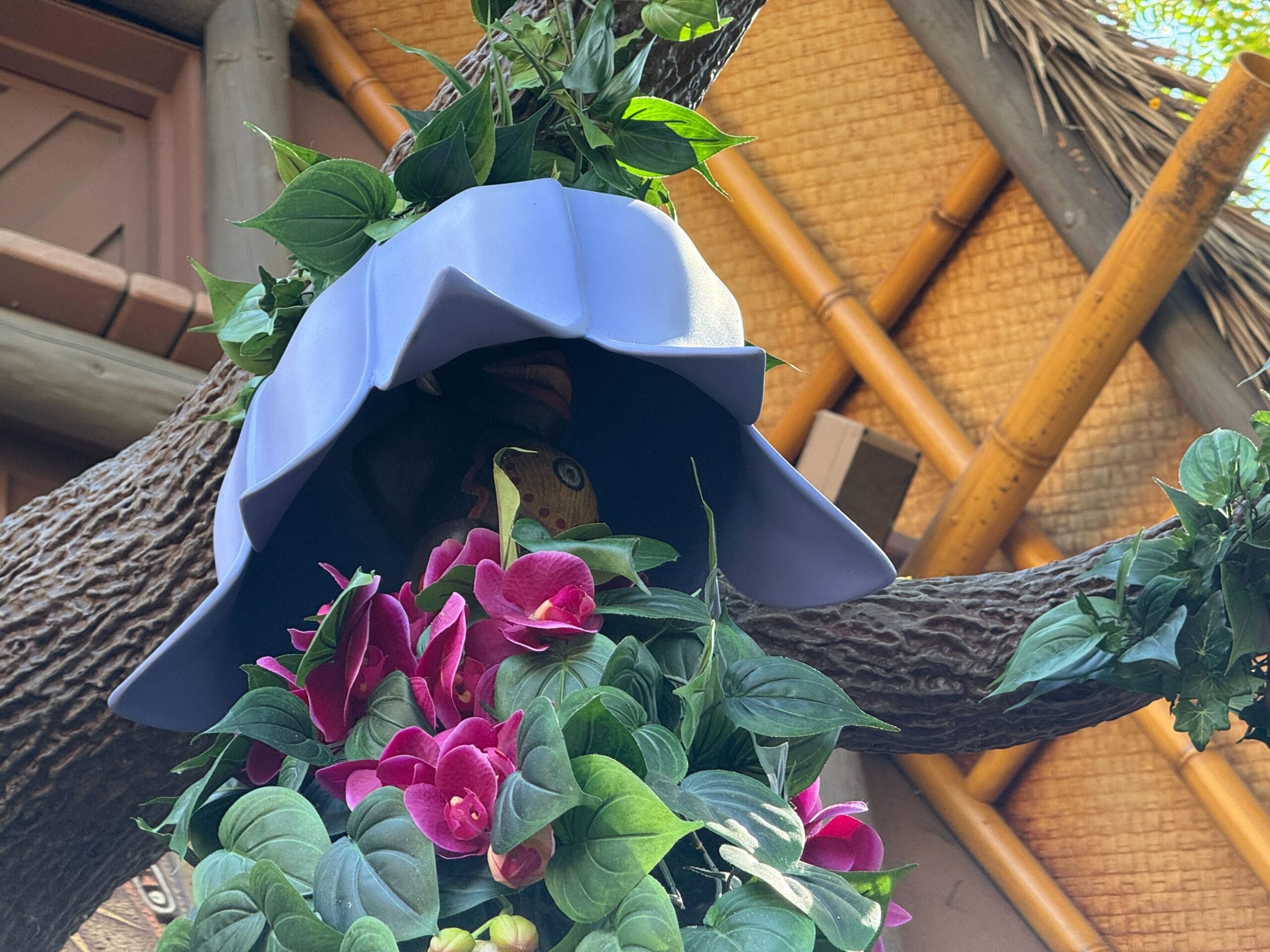 A plastic bird peeks from beneath a blue hat amid green leaves and pink flowers in the Enchanted Tiki Garden.