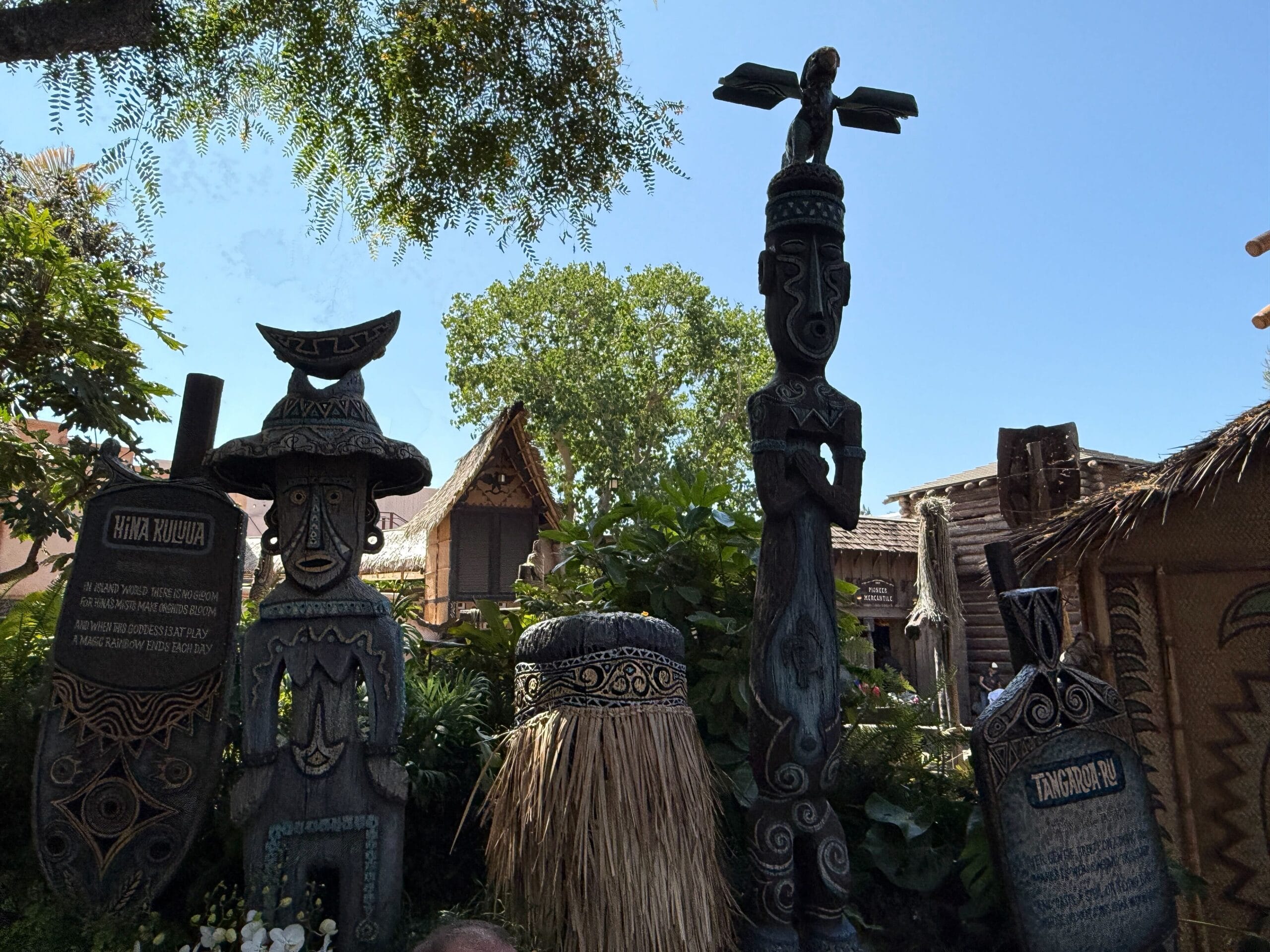Three carved tiki statues stand in the Enchanted Tiki Garden, lush with plants and a thatched-roof hut under blue skies.