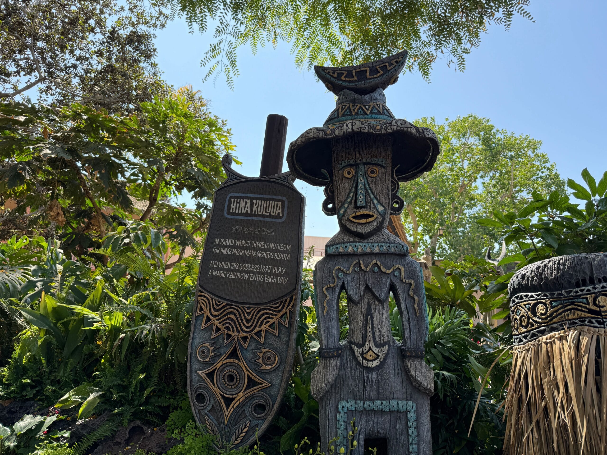 A wooden shield and tiki statue with carvings stand in the lush Enchanted Tiki Garden under a sunny sky.