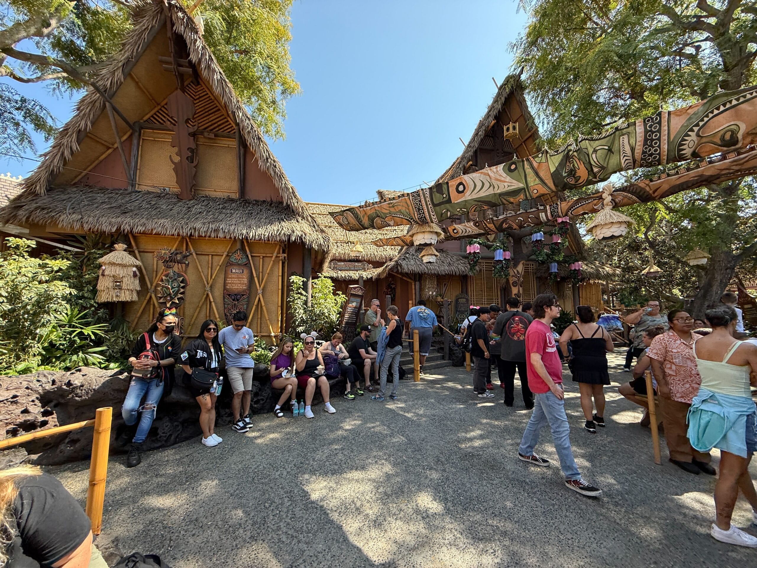 People gather outside Walt Disney’s Enchanted Tiki Room, near thatched roofs and banners under a sunny sky.