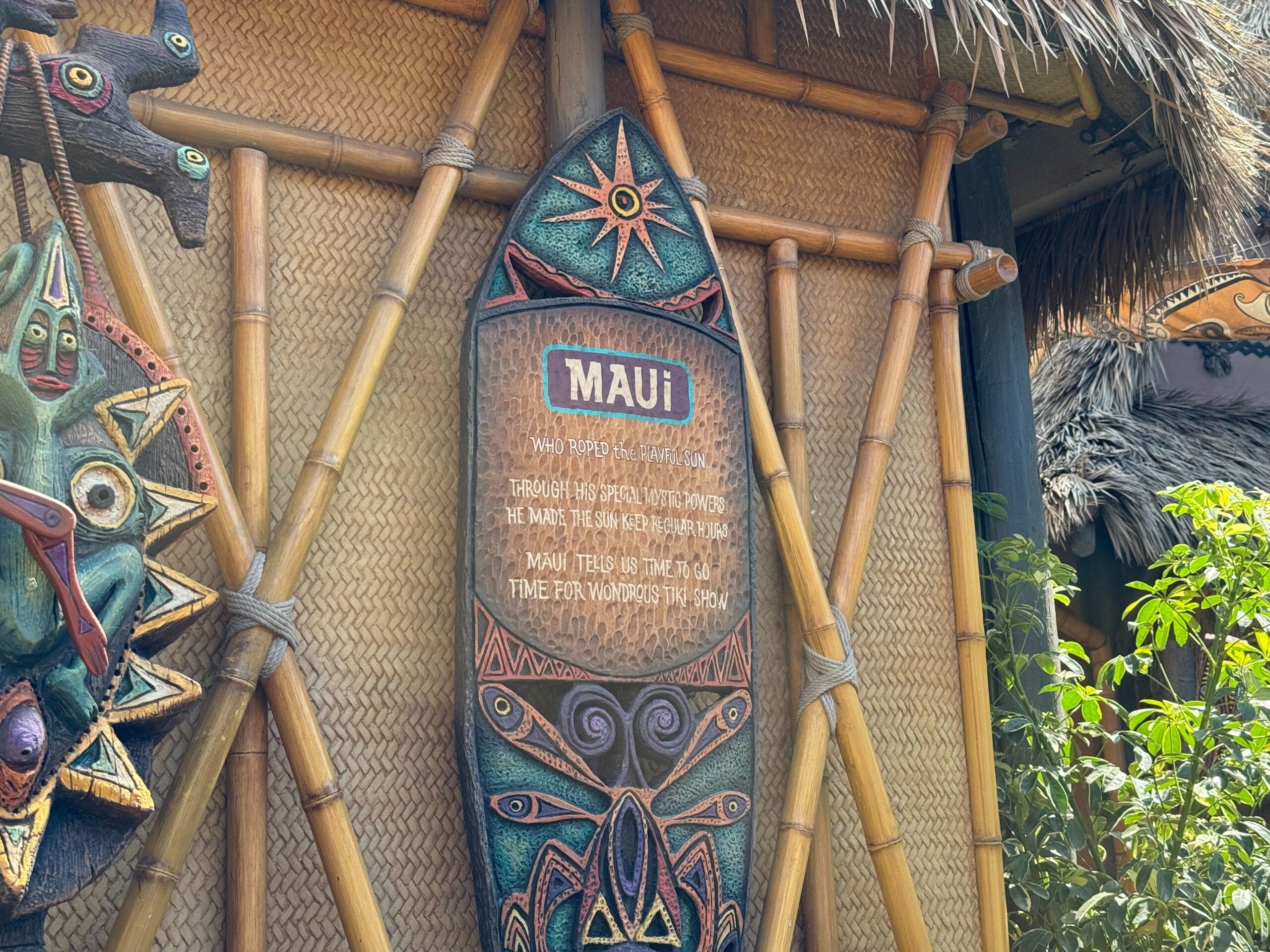 A decorative wooden Maui sign in the Enchanted Tiki Garden, mounted on bamboo and surrounded by tropical plants.