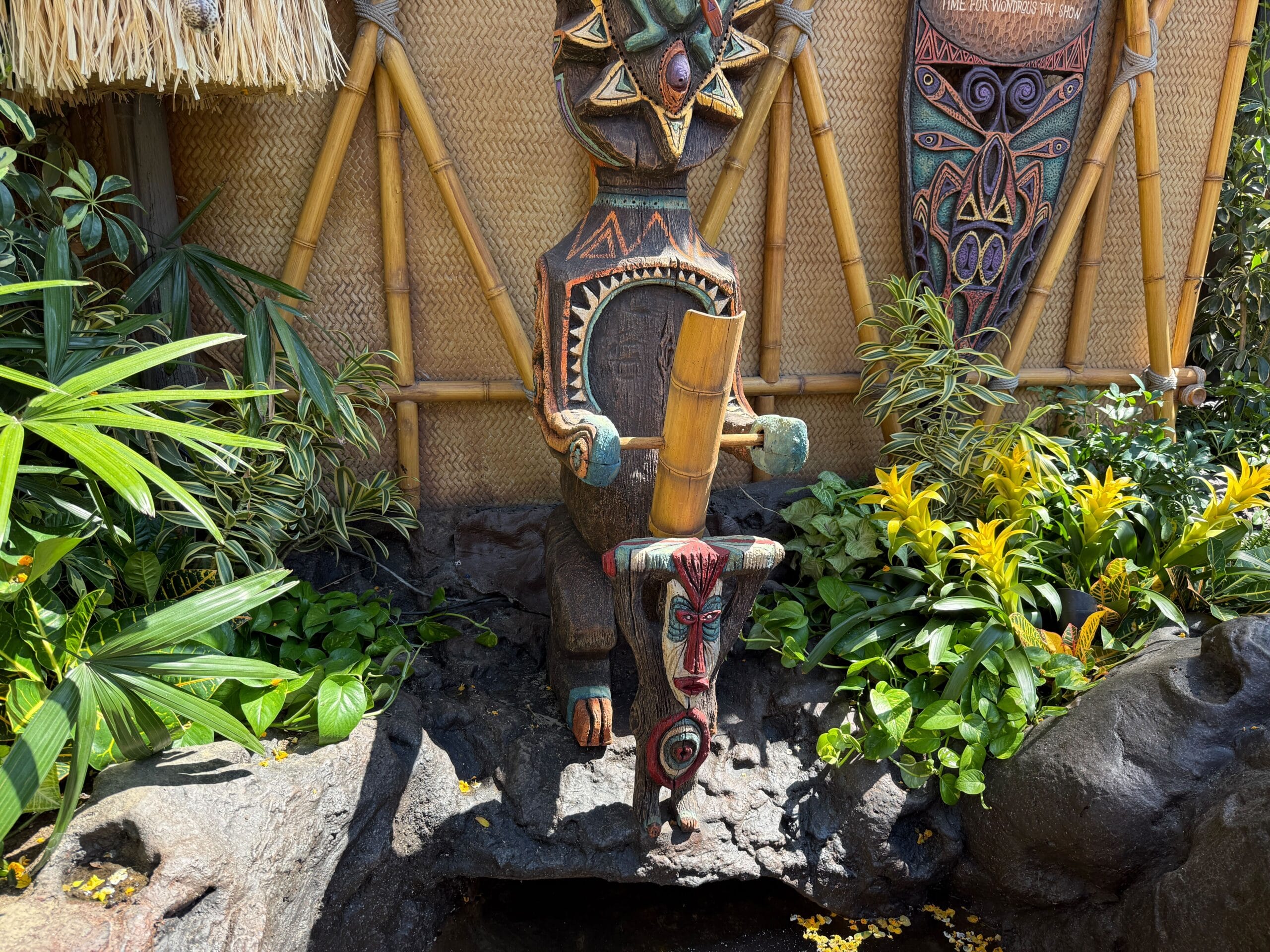 A colorful tiki statue with a bamboo tube stands in the Enchanted Tiki Garden amid lush plants and rocks.