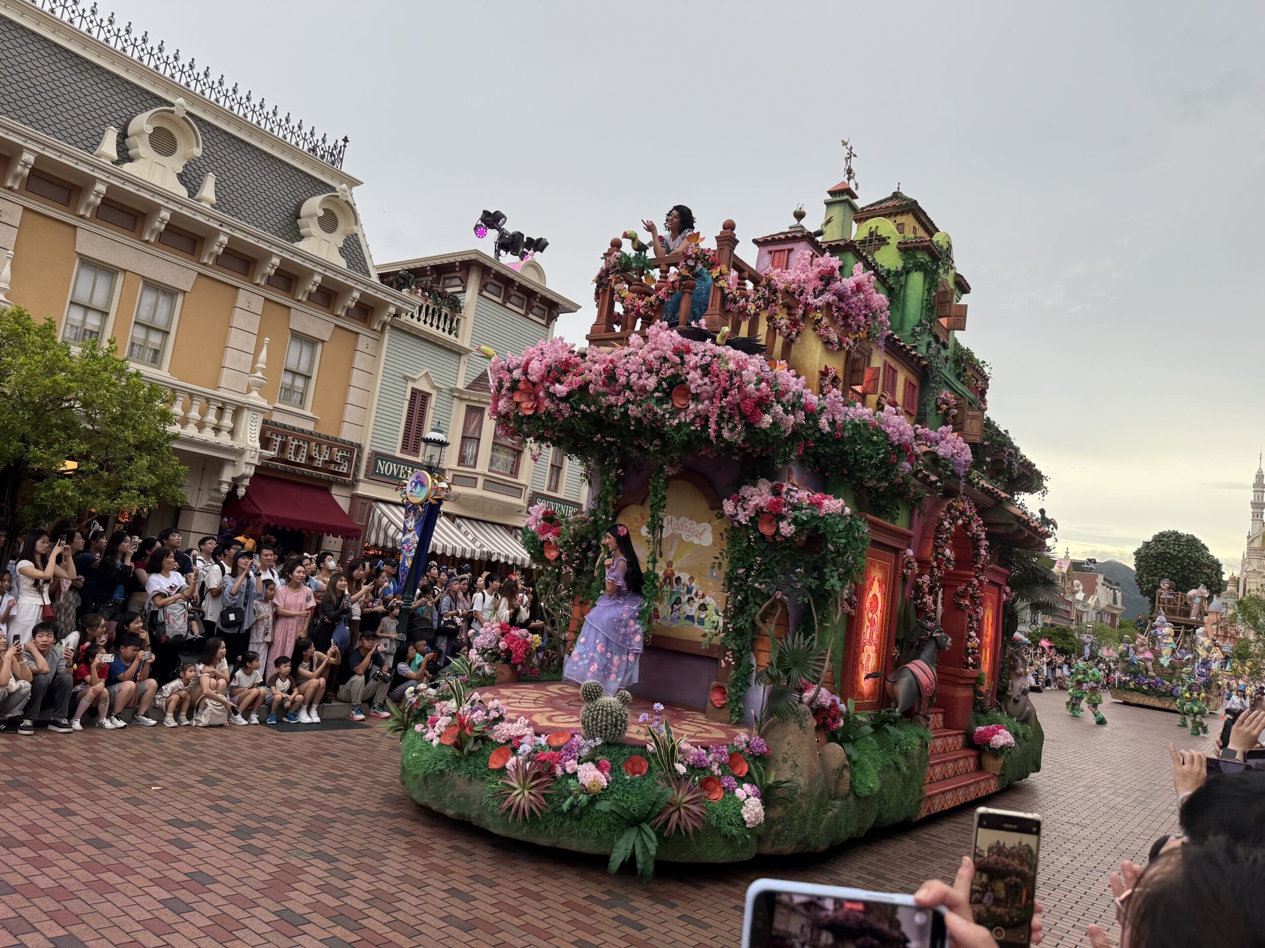 A colorful Friendtastic Parade float with flowers and characters passes a crowd at Hong Kong Disneyland as people take photos.