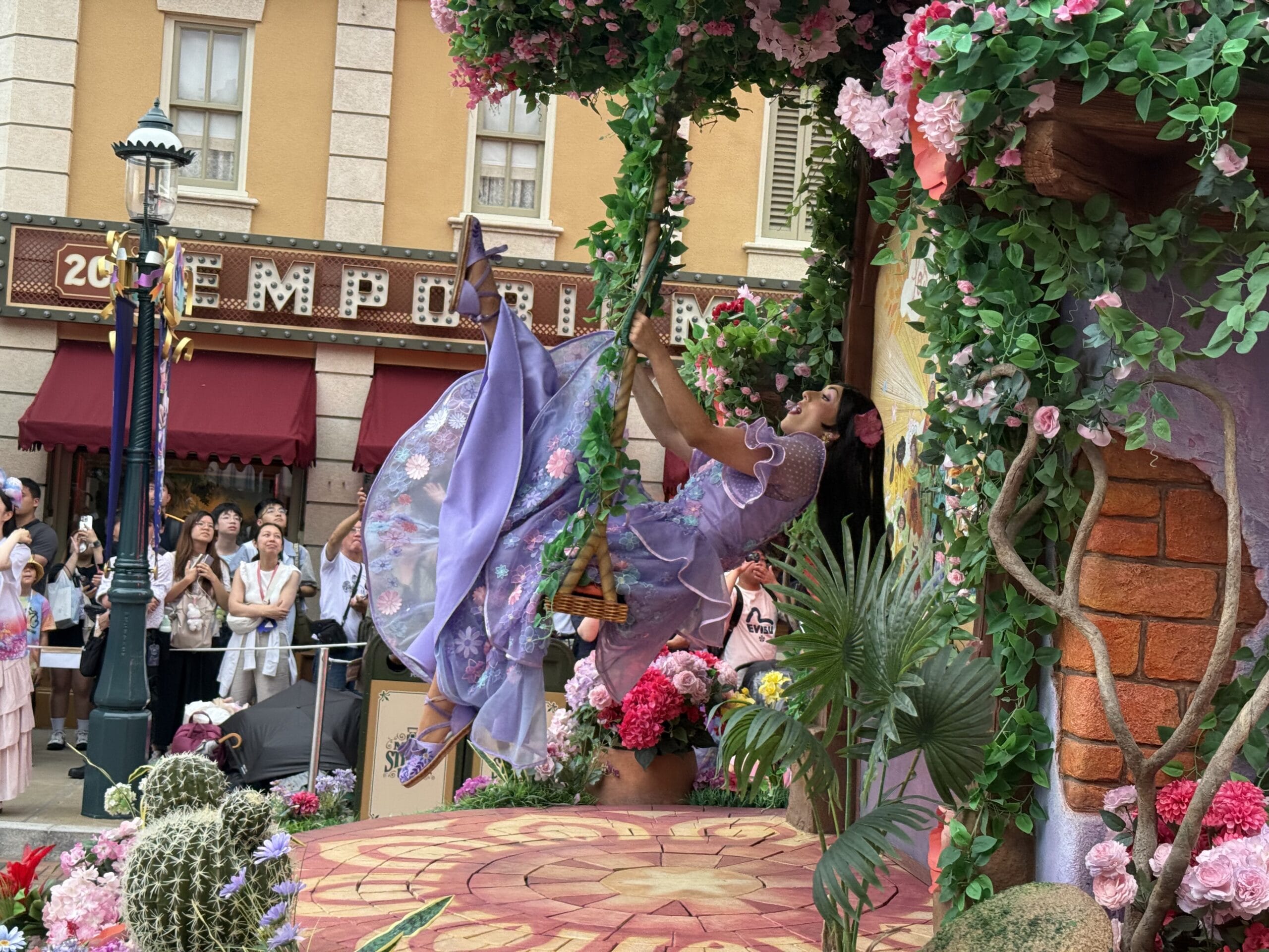 A woman in a purple dress swings on a vine at the Friendtastic Parade, with colorful flowers and crowds at Hong Kong Disneyland.