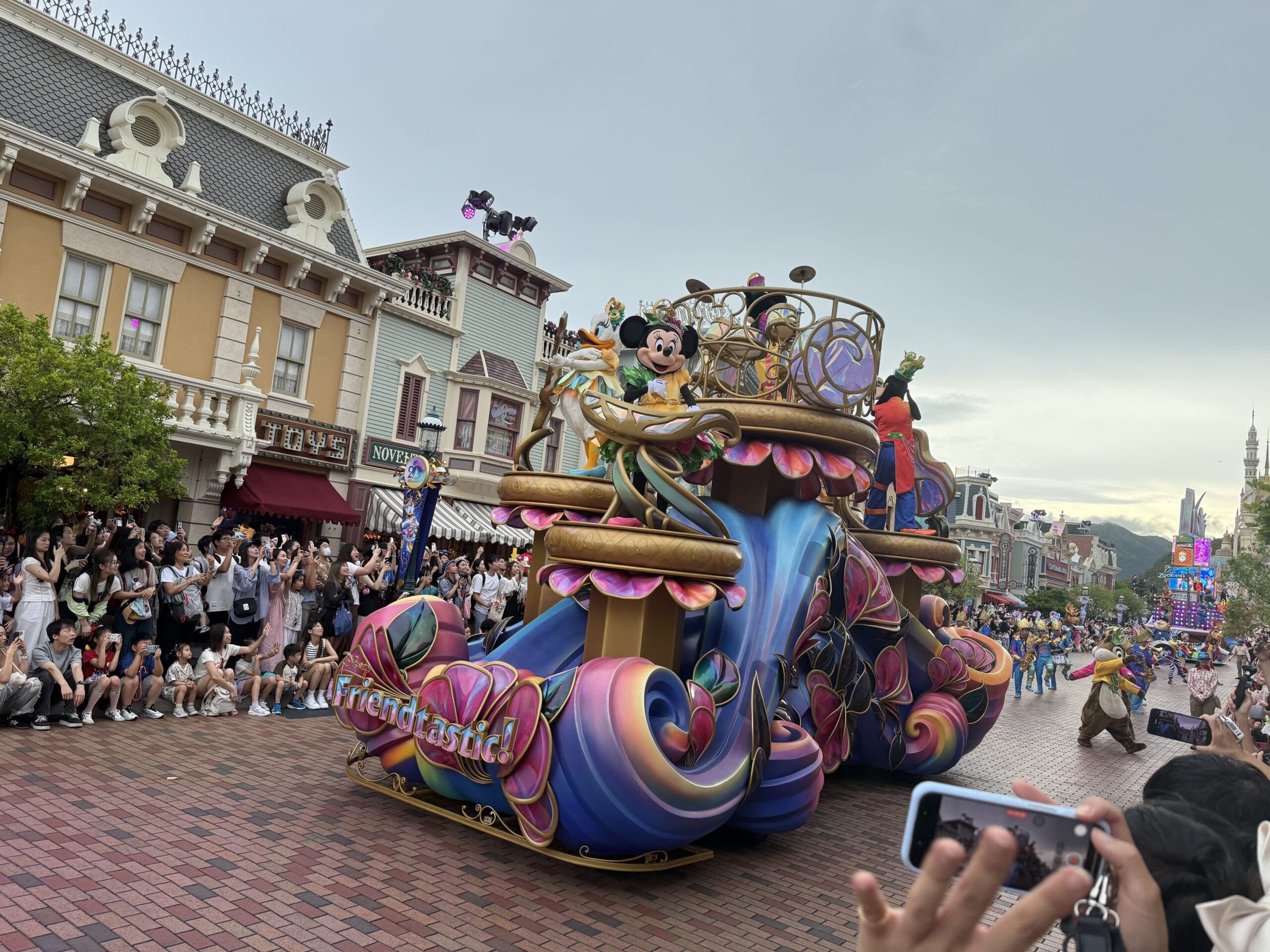 Colorful friendtastic parade float with Mickey Mouse passes crowds at Hong Kong Disneyland as people snap photos.
