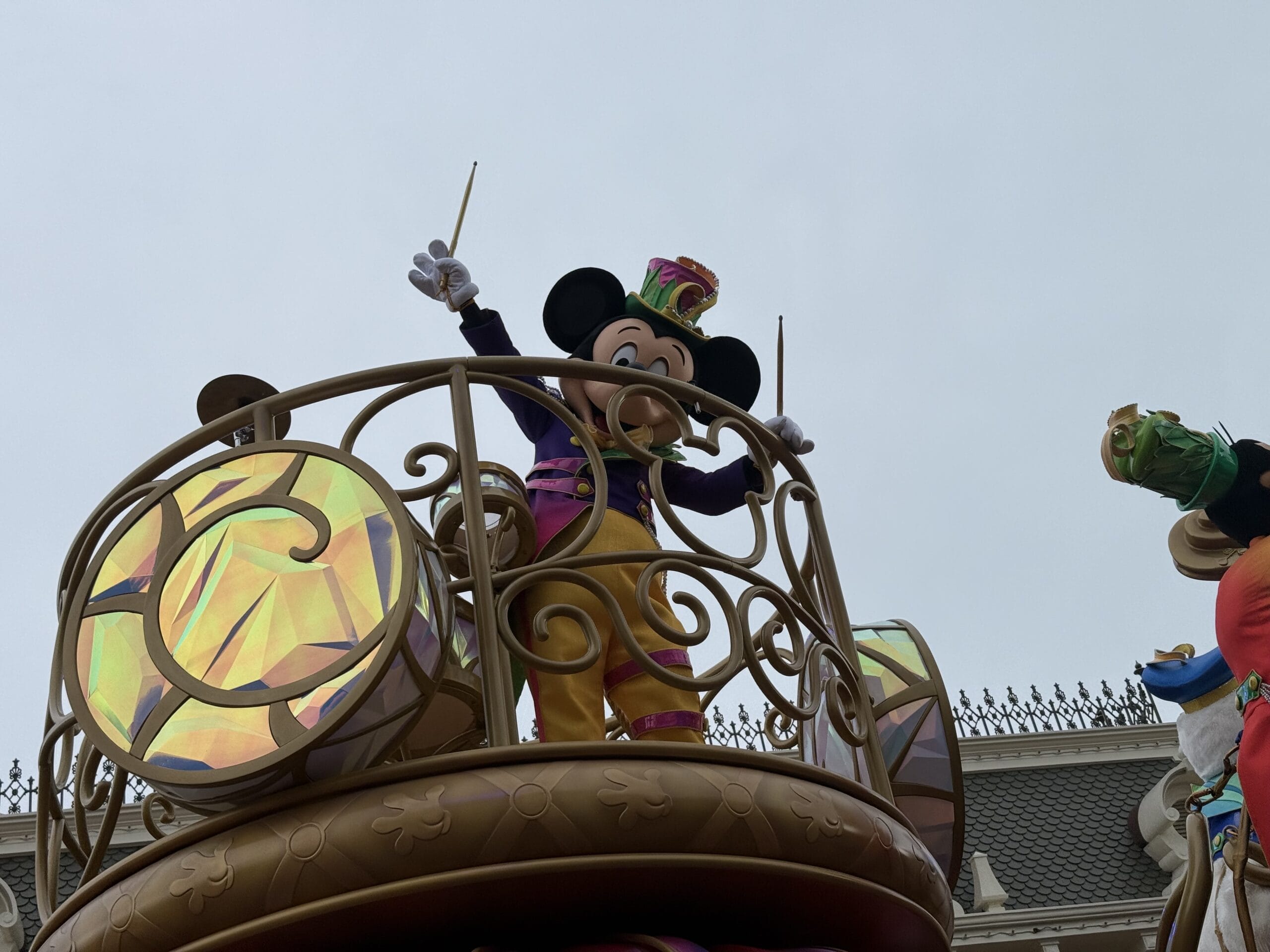 Mickey Mouse in a colorful outfit conducts on the Hong Kong Disneyland Friendtastic Parade float with shiny drums and railings.