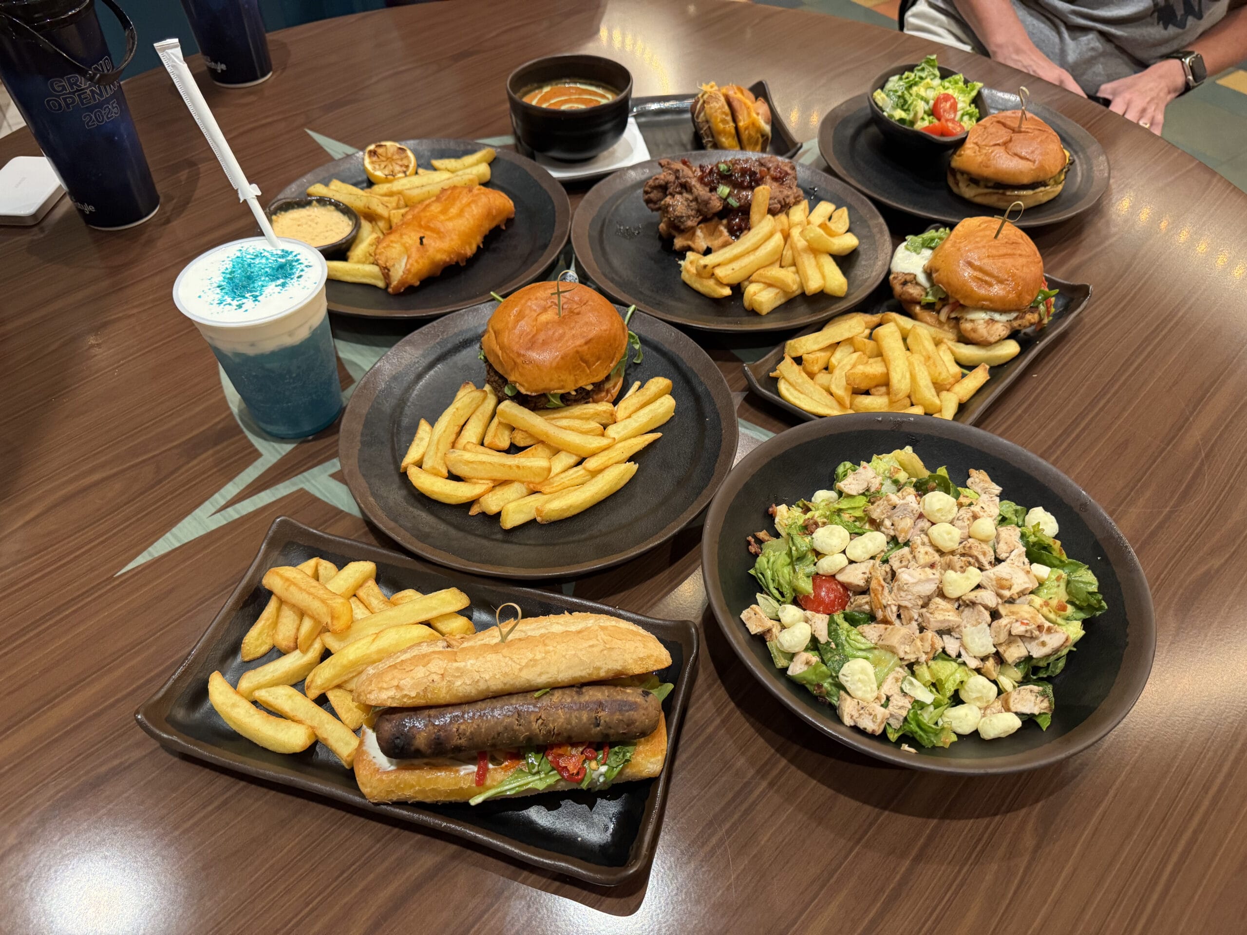 A Meteor Astropub table in Epic Universe with burgers, fries, salad, sausage sandwich, fish and chips, and blue drink.