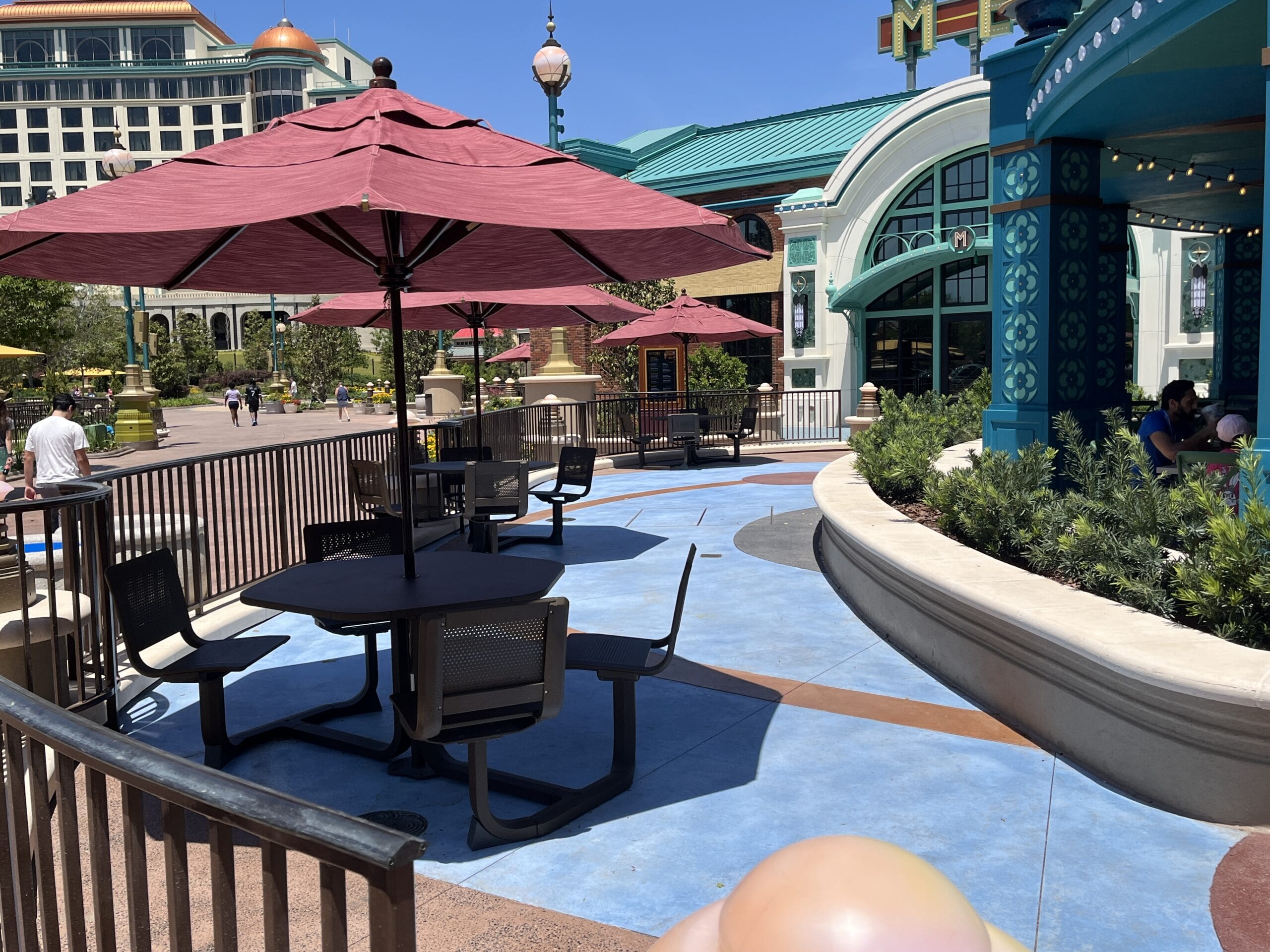 Outdoor patio at Meteor Astropub with red umbrellas, near Epic Universe’s colorful buildings on a sunny day.