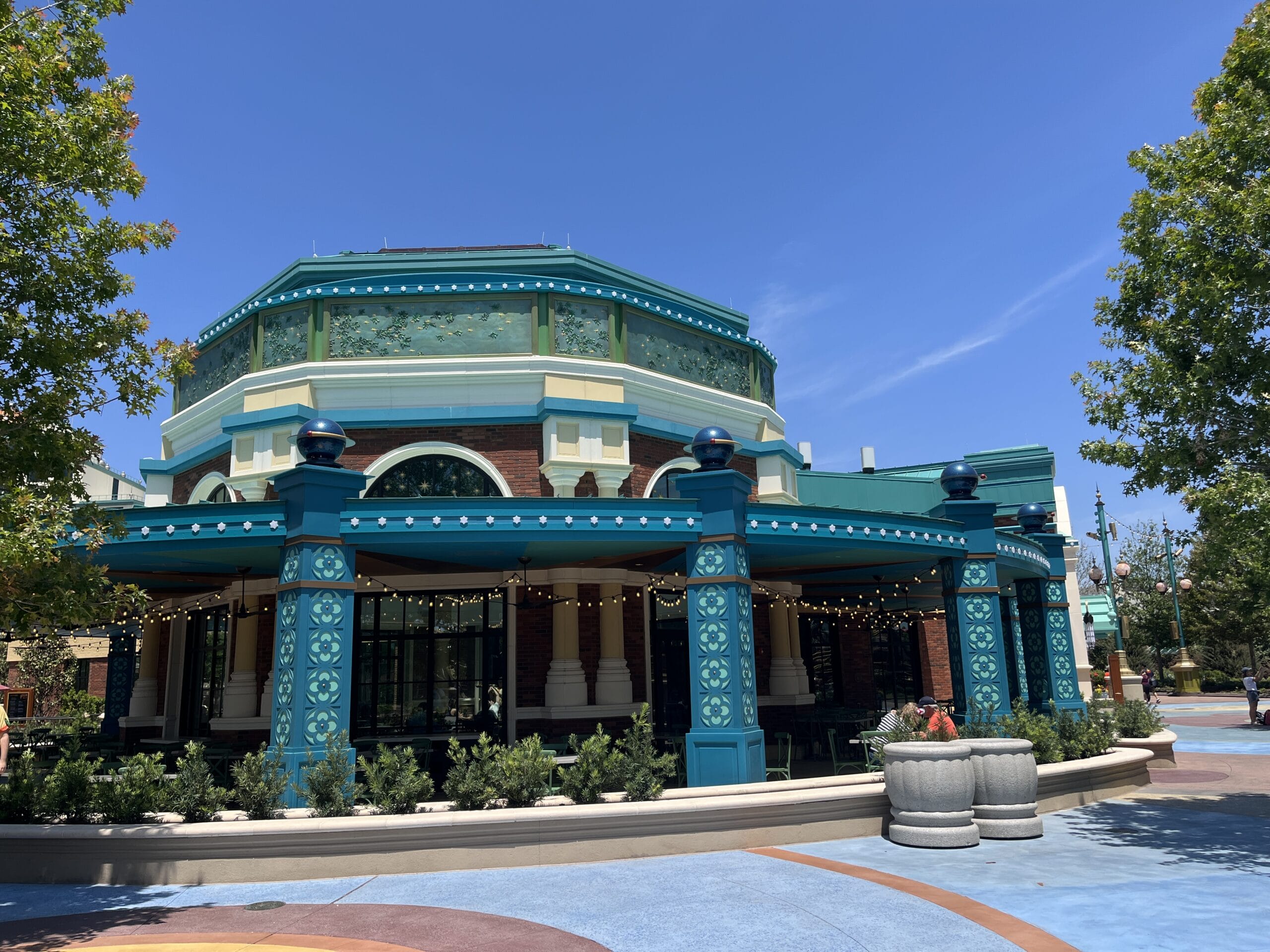 A colorful round building with teal pillars at Meteor Astropub, string lights, and trees on a sunny Epic Universe day.