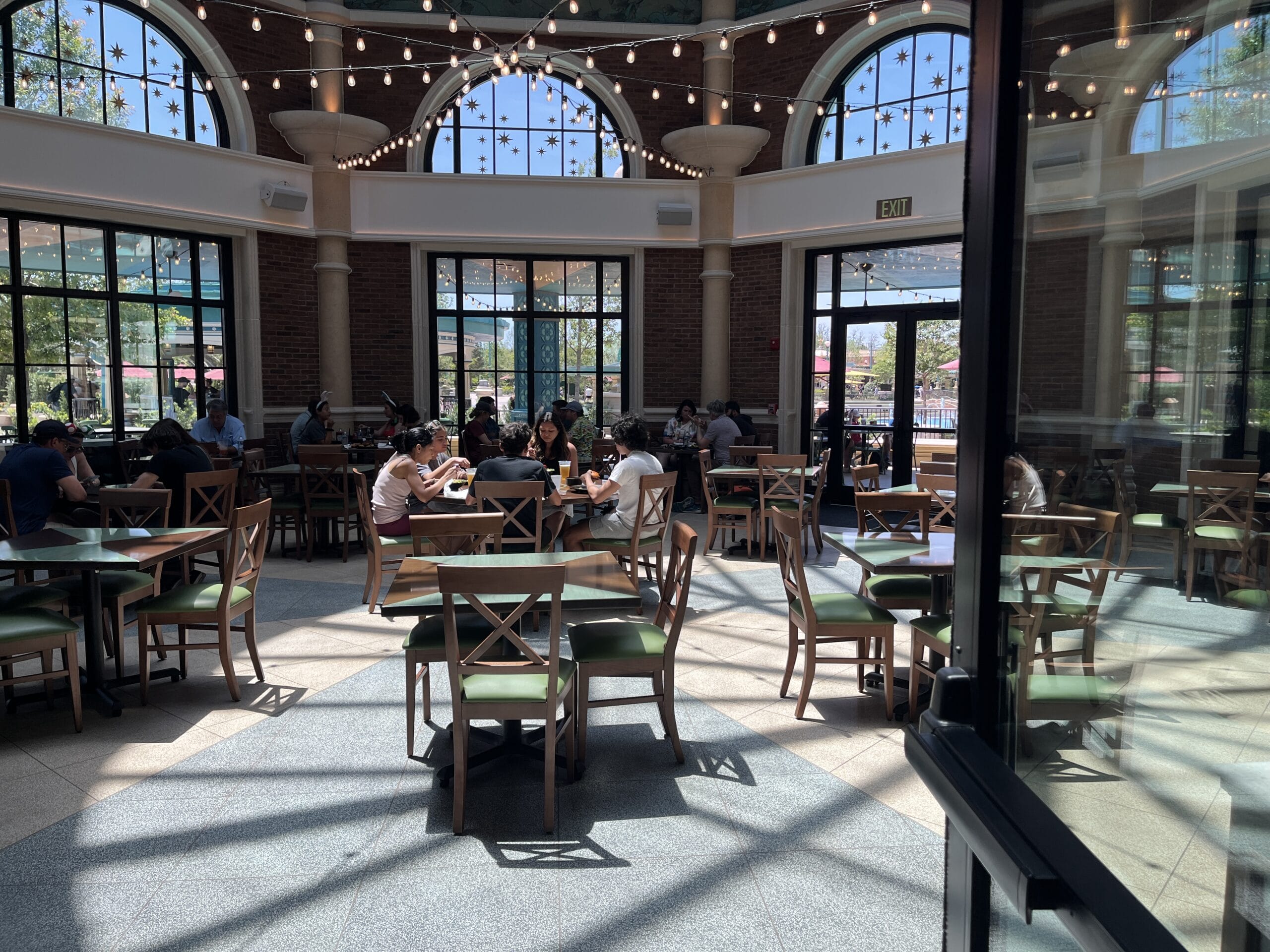 People dine at wooden tables in Meteor Astropub, a sunlit, glass-walled restaurant with string lights and high ceilings.