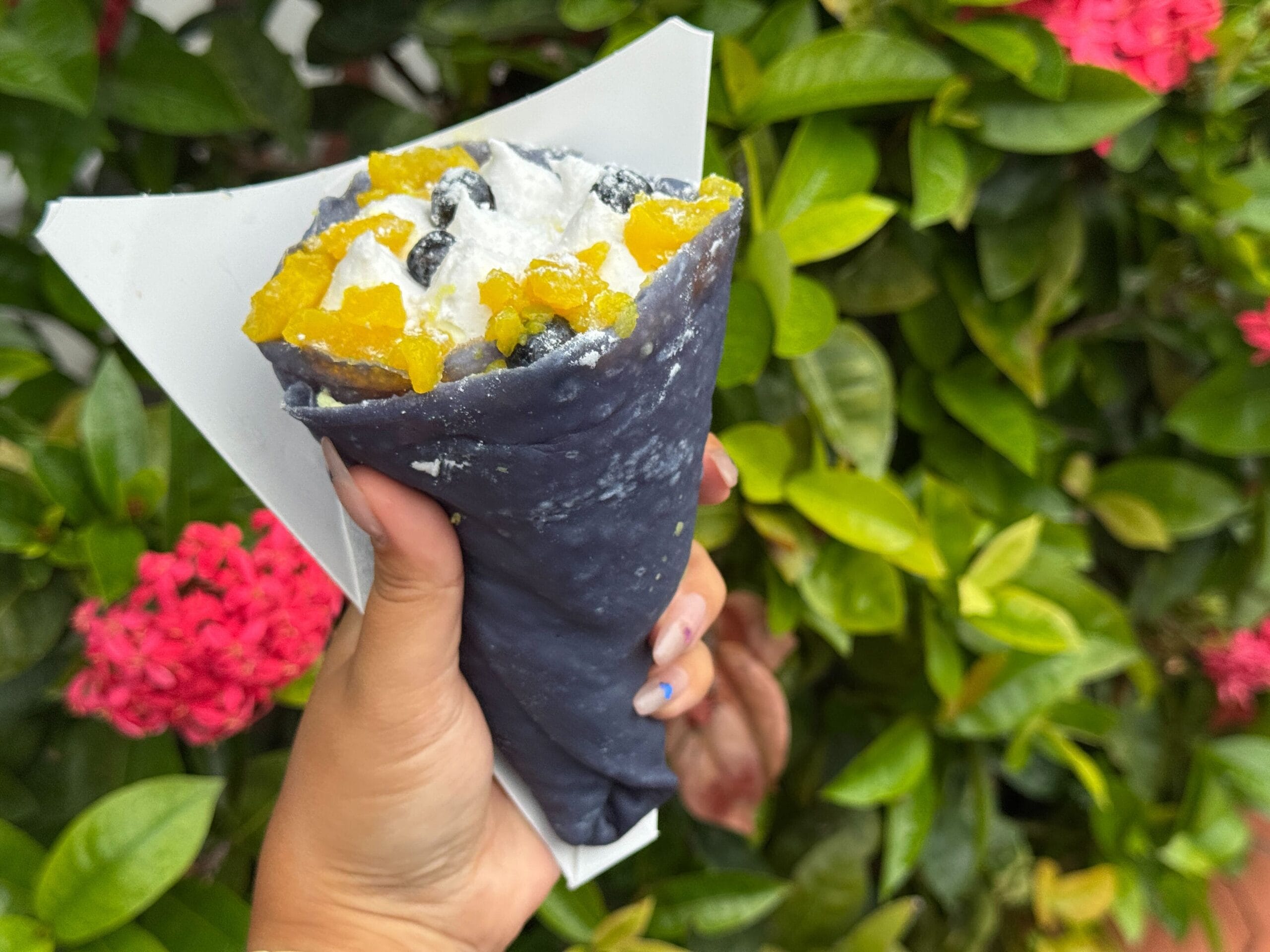 A hand holds a blue crepe filled with cream, yellow fruit, and boba pearls, with green leaves and pink flowers in the background.