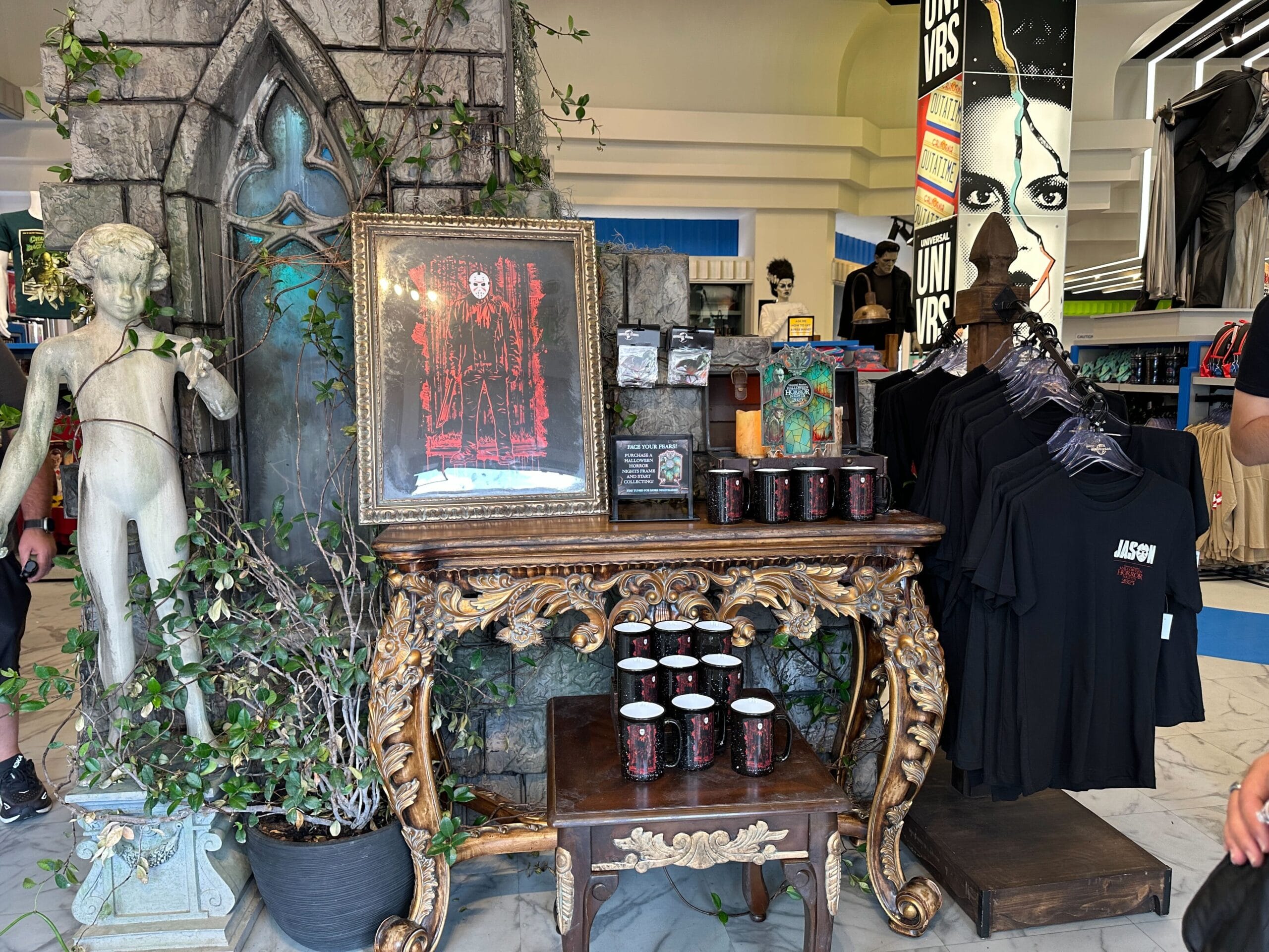 A gothic-themed display with horror mugs, "Jason" shirts, Friday the 13th art, a statue, and decorative plants.