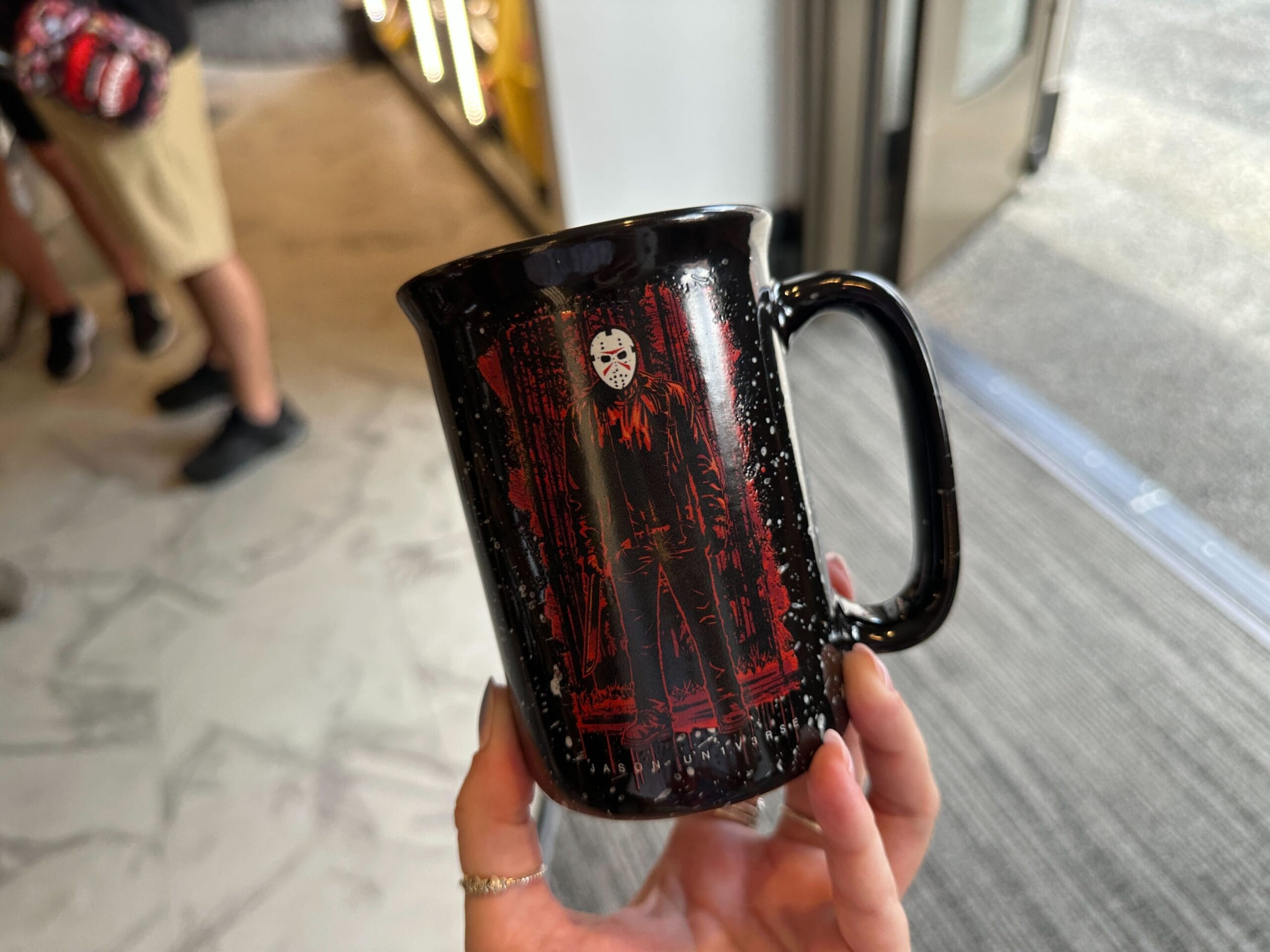 A hand holds a black mug with a red and white illustration from the Jason Universe: Friday the 13th.