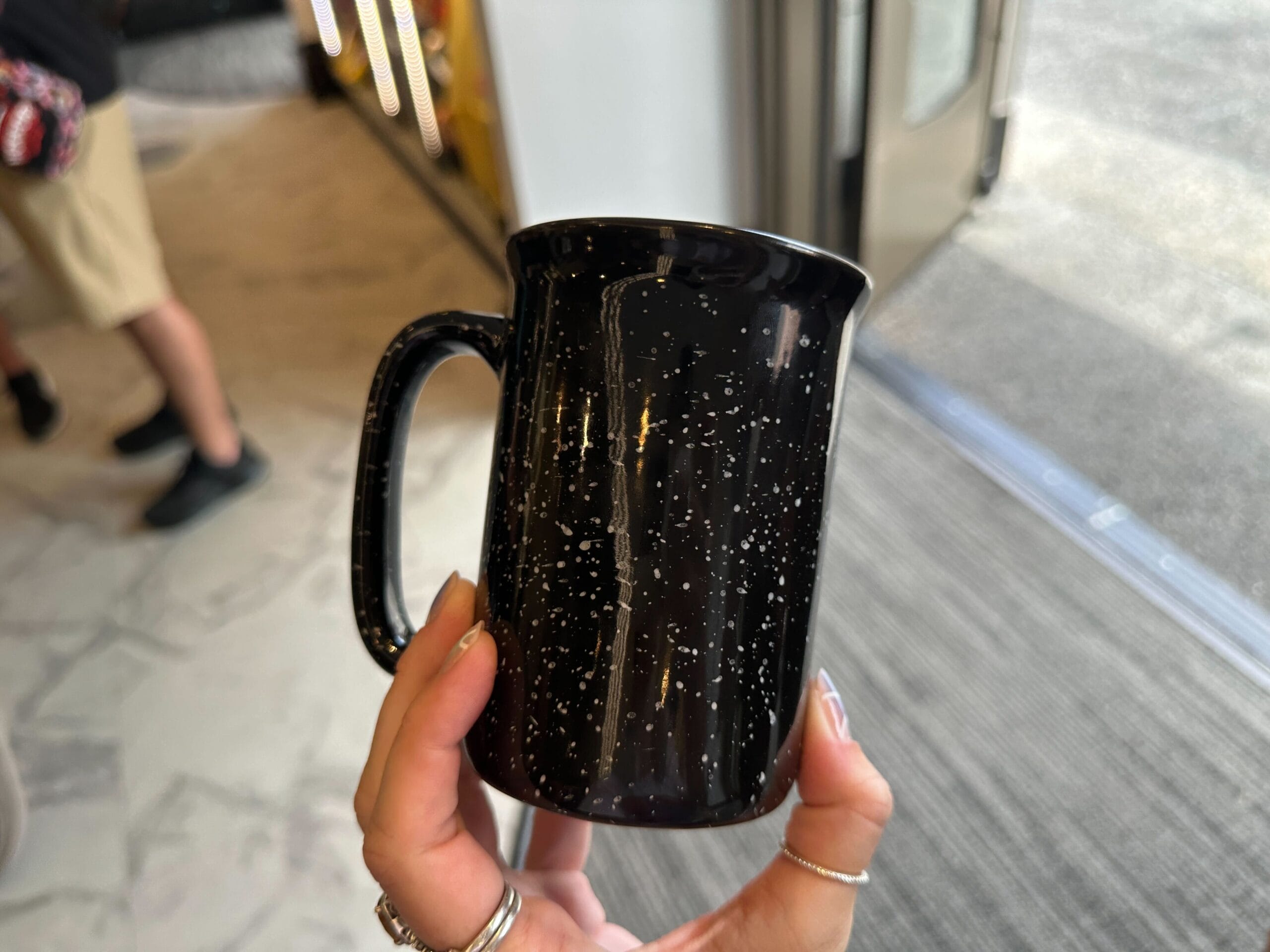 A hand holds a black mug with white speckles, resembling the Jason Universe, indoors near a glass door.