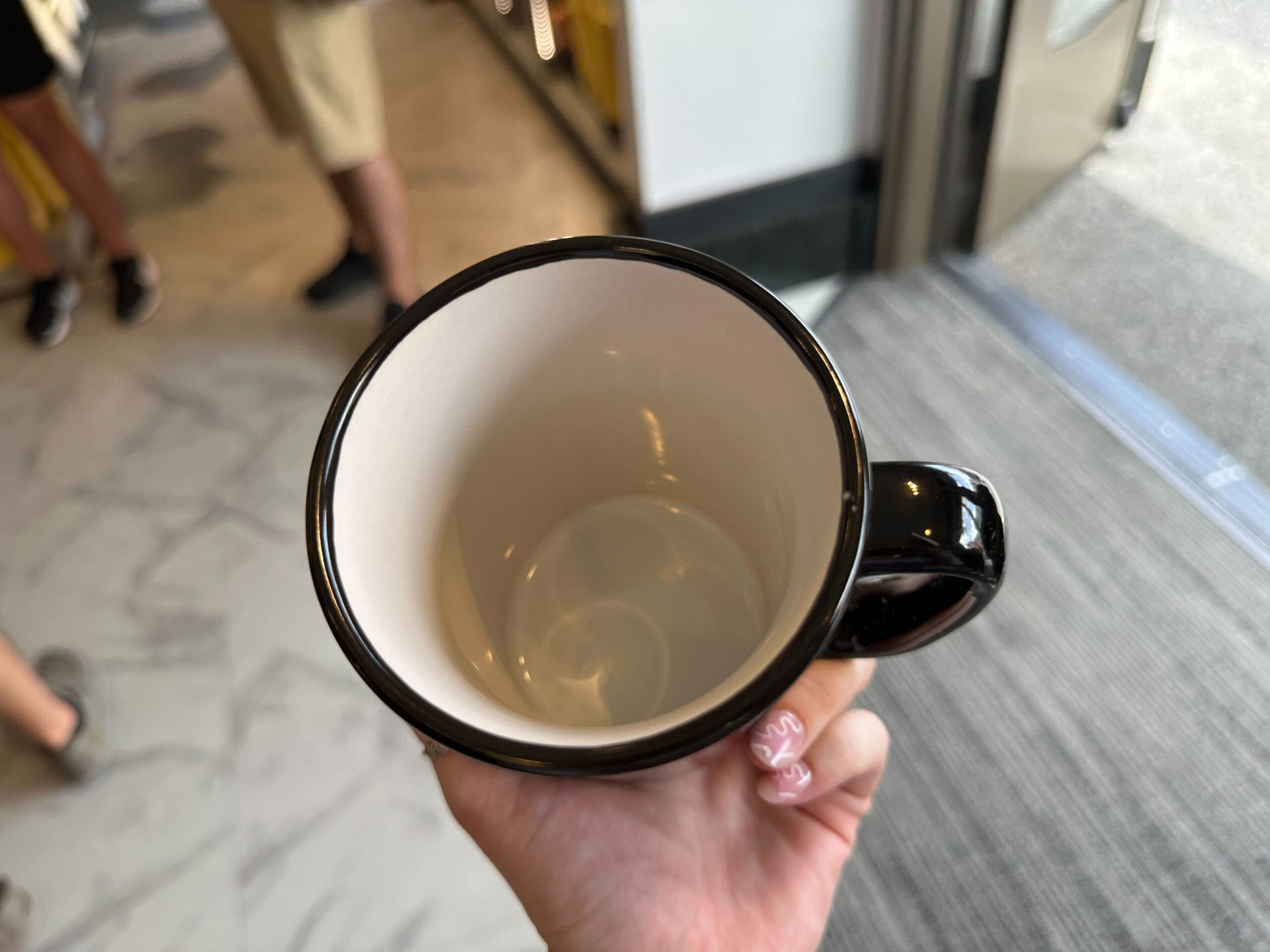 Hand holding an empty ceramic mug in a store at Halloween Horror Nights 34, people and flooring in the background.