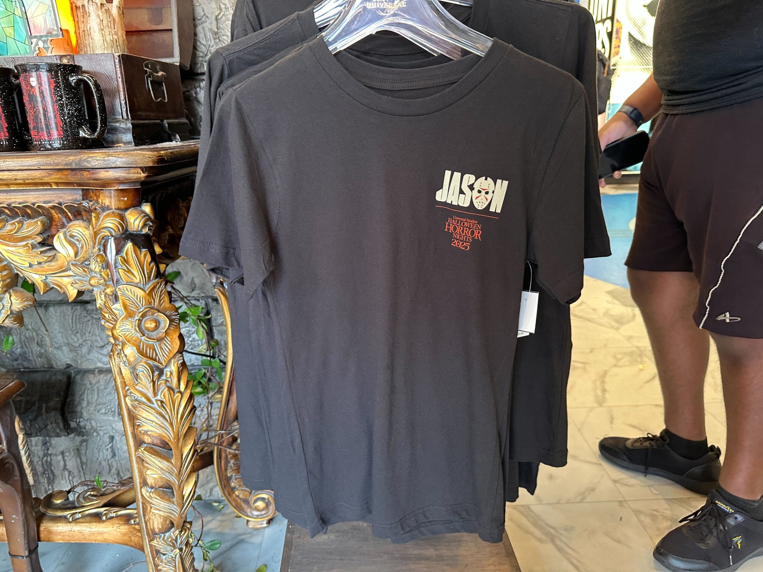 A black T-shirt on a hanger with "JASON" and "Halloween Horror Nights 2023" from the Friday the 13th universe printed on front.