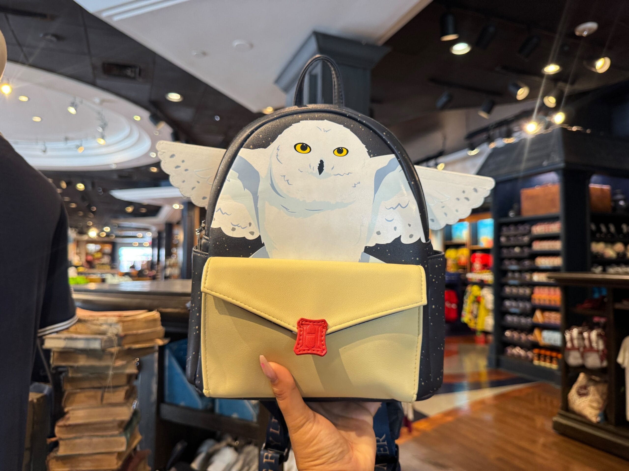 A hand holds a Harry Potter backpack with a Hedwig design in a store with shelves and lights in the background.