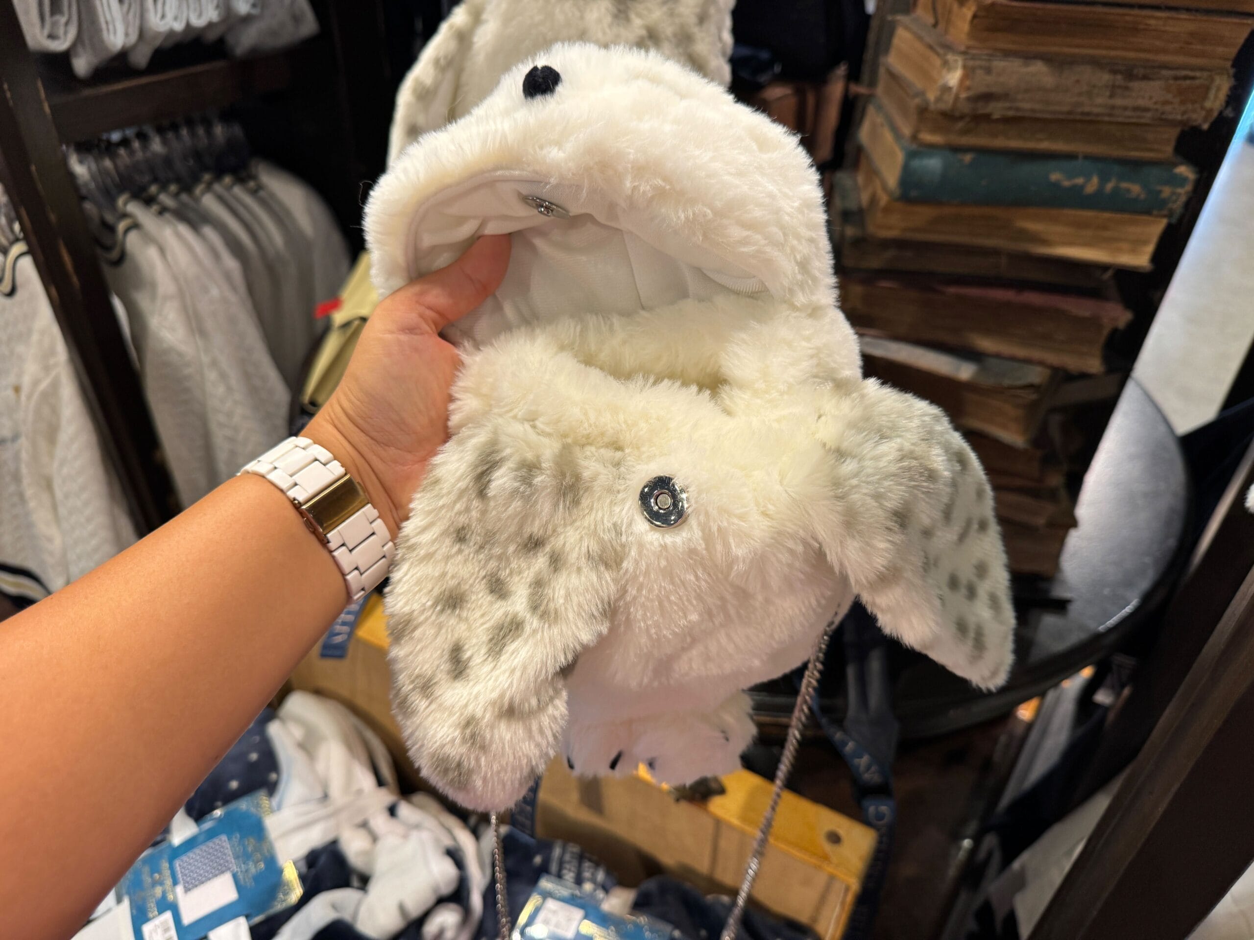 A hand holds a plush white Snow Owl Plush purse with gray spots and button details in a store.