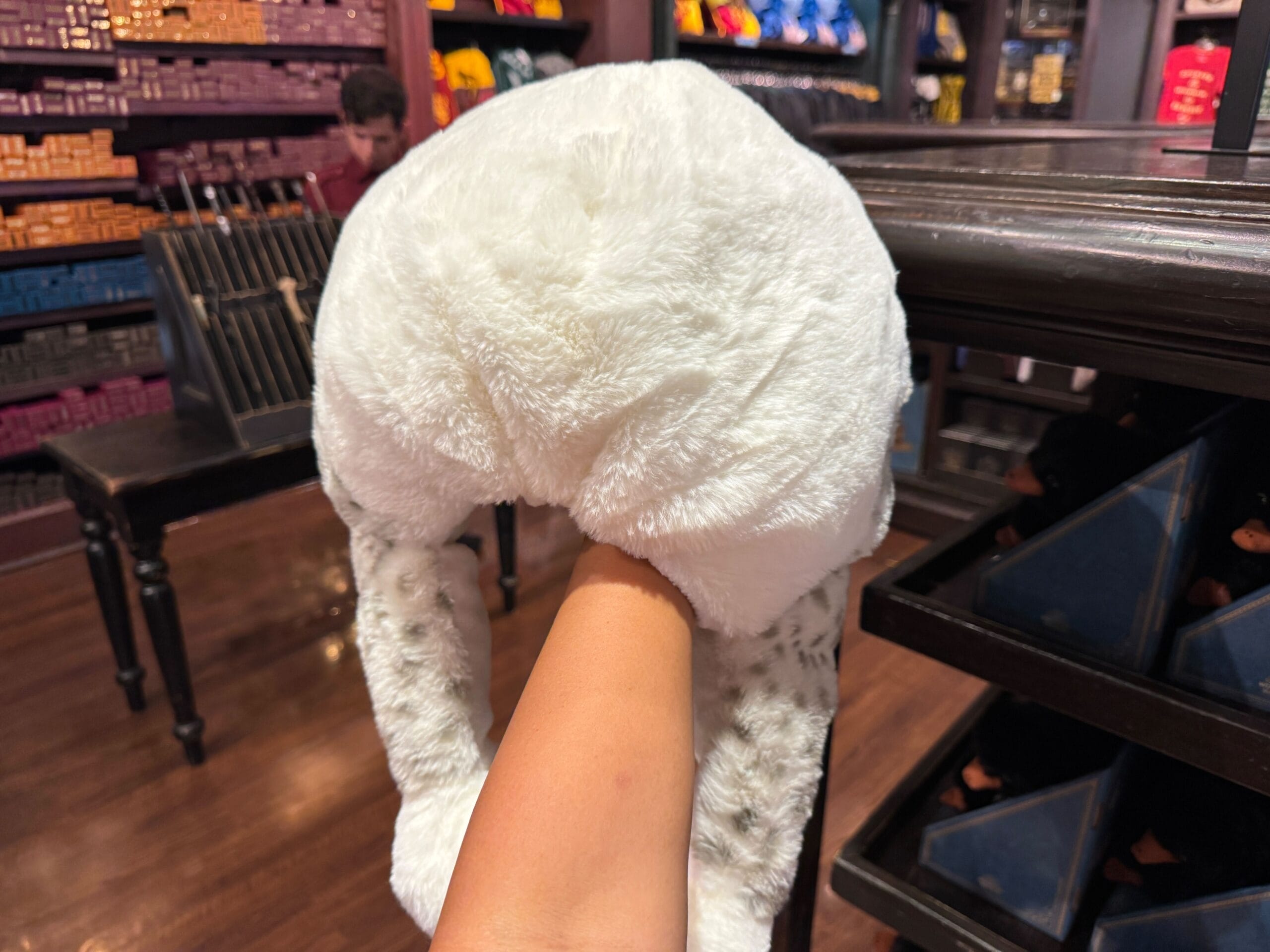 A hand holds a white Owl Hat with ear flaps in a store filled with colorful merchandise on shelves.