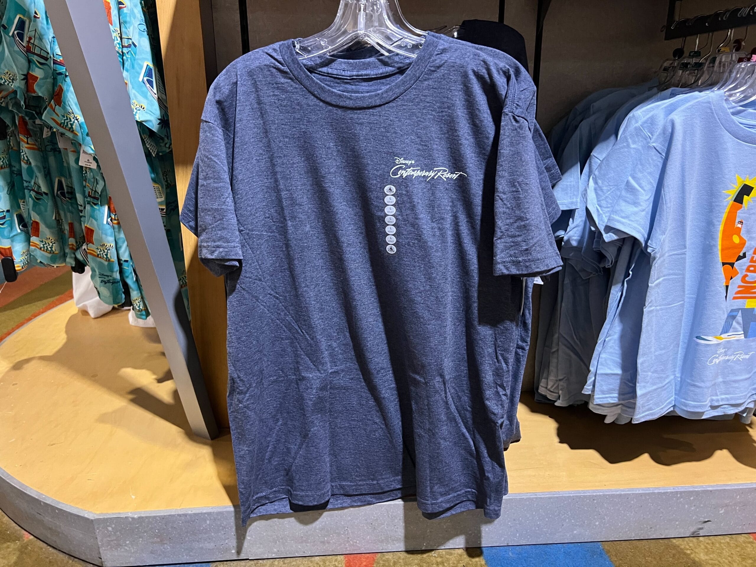 A blue Grand Floridian Resort t-shirt hangs on a rack beside Contemporary resort tees in the store.