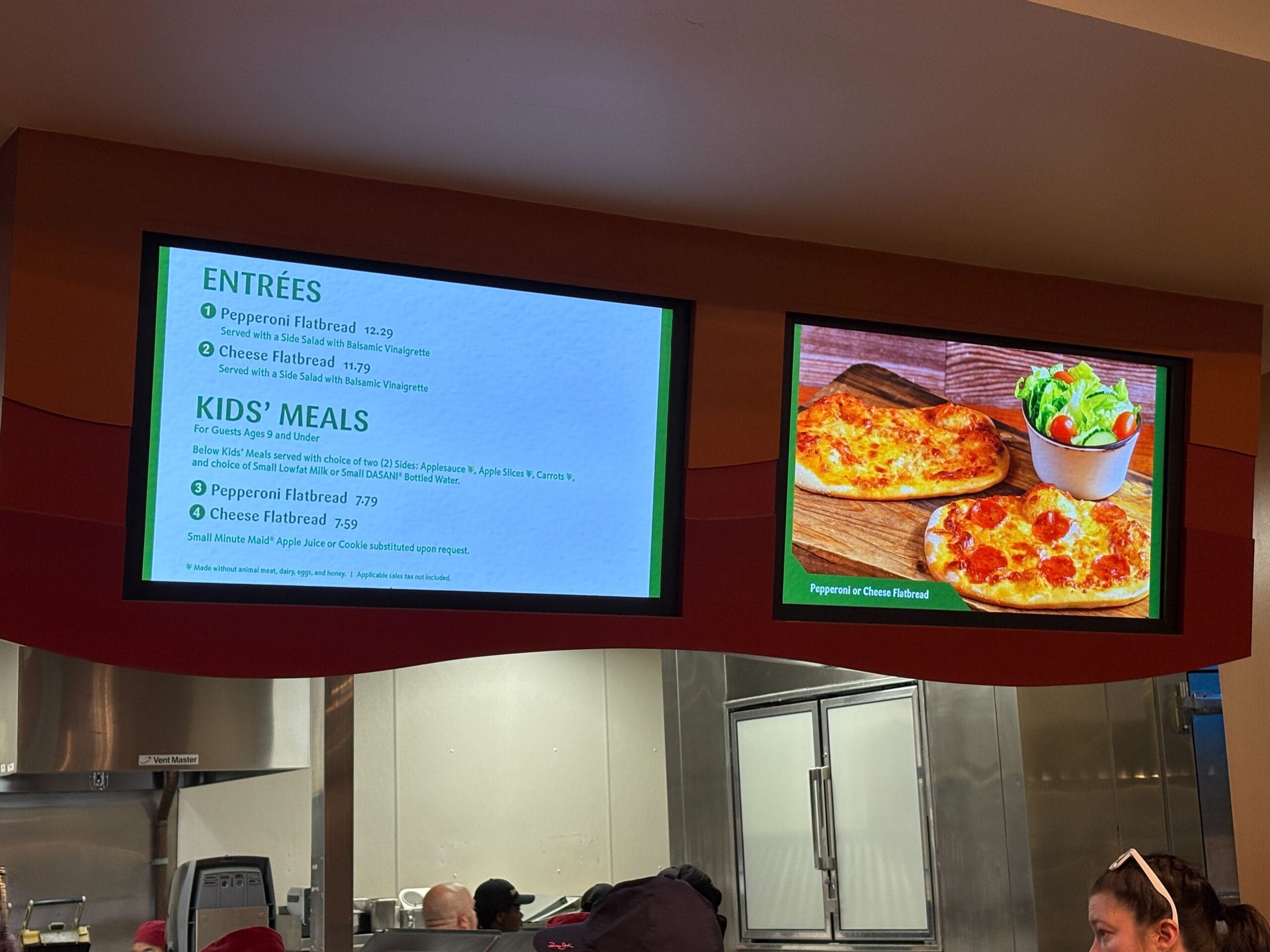 Digital menu at Sunshine Seasons displays pizza, flatbreads, salads, and kids’ meals above a visible kitchen counter.
