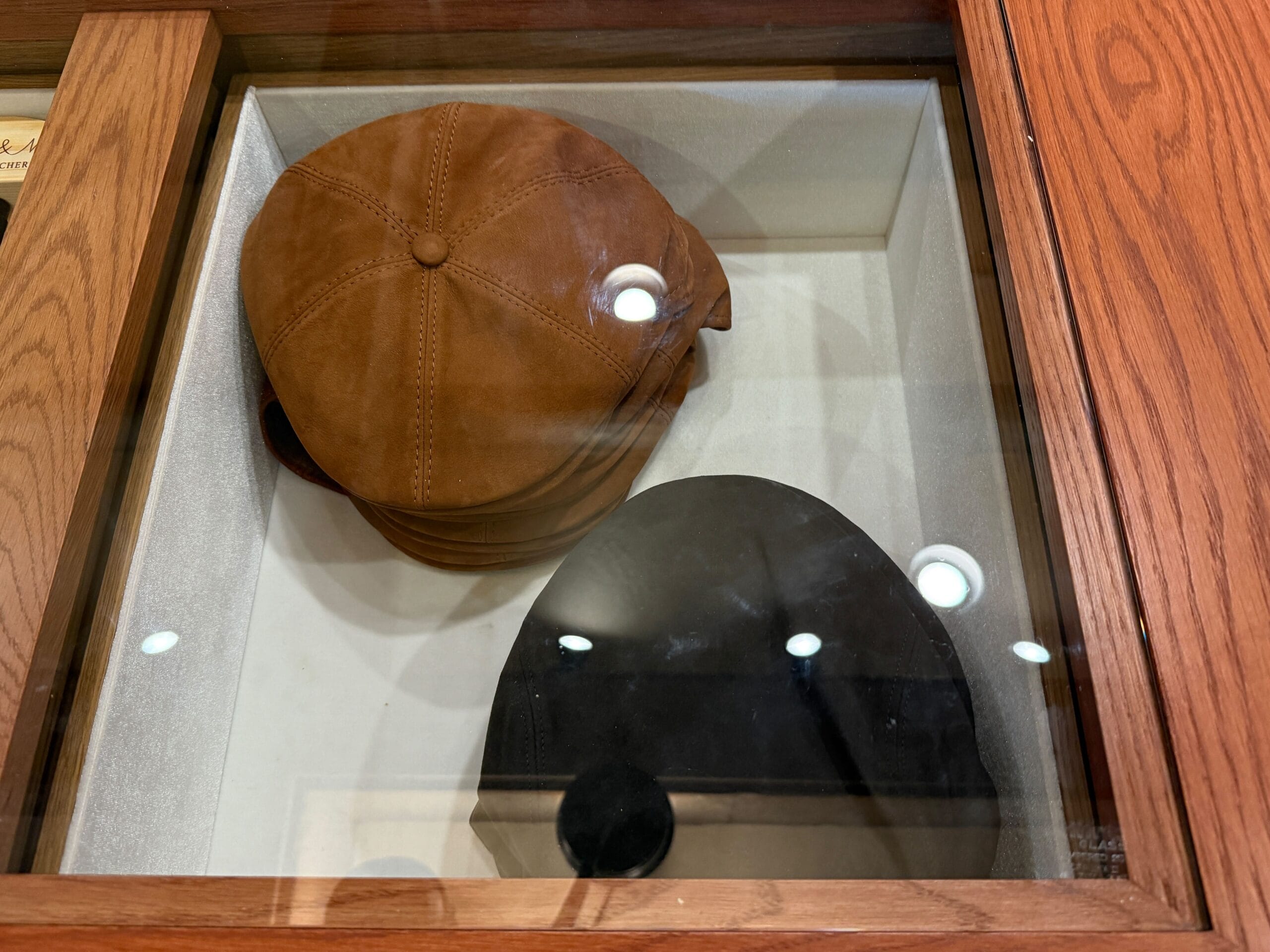 Two flat caps from the Prairie Outpost Hat Shop, brown and black, displayed under glass with overhead light reflections.
