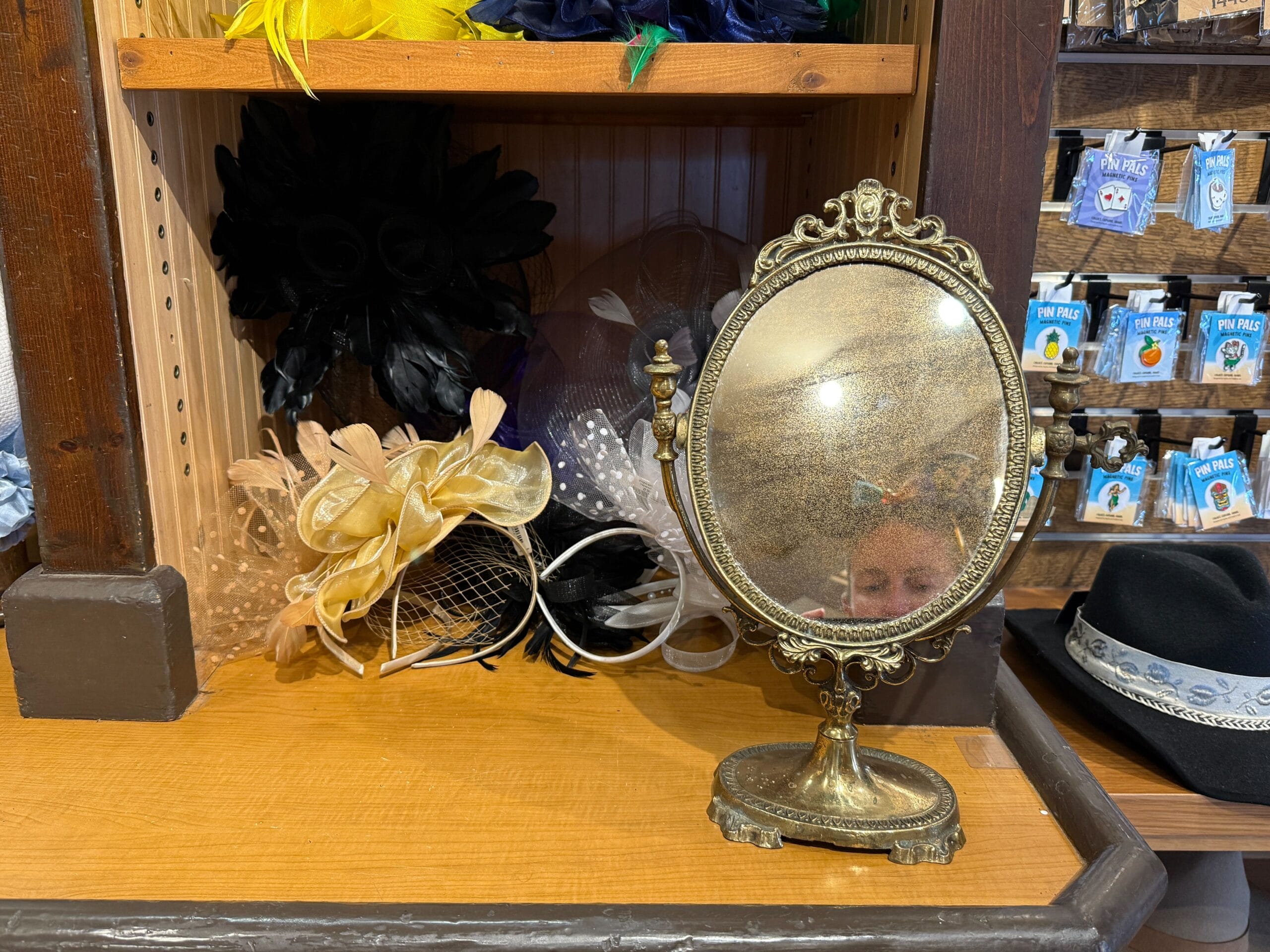 Vintage oval mirror on a wooden shelf at the Hat Shop, with fascinators and hats in the background; partial reflection visible.