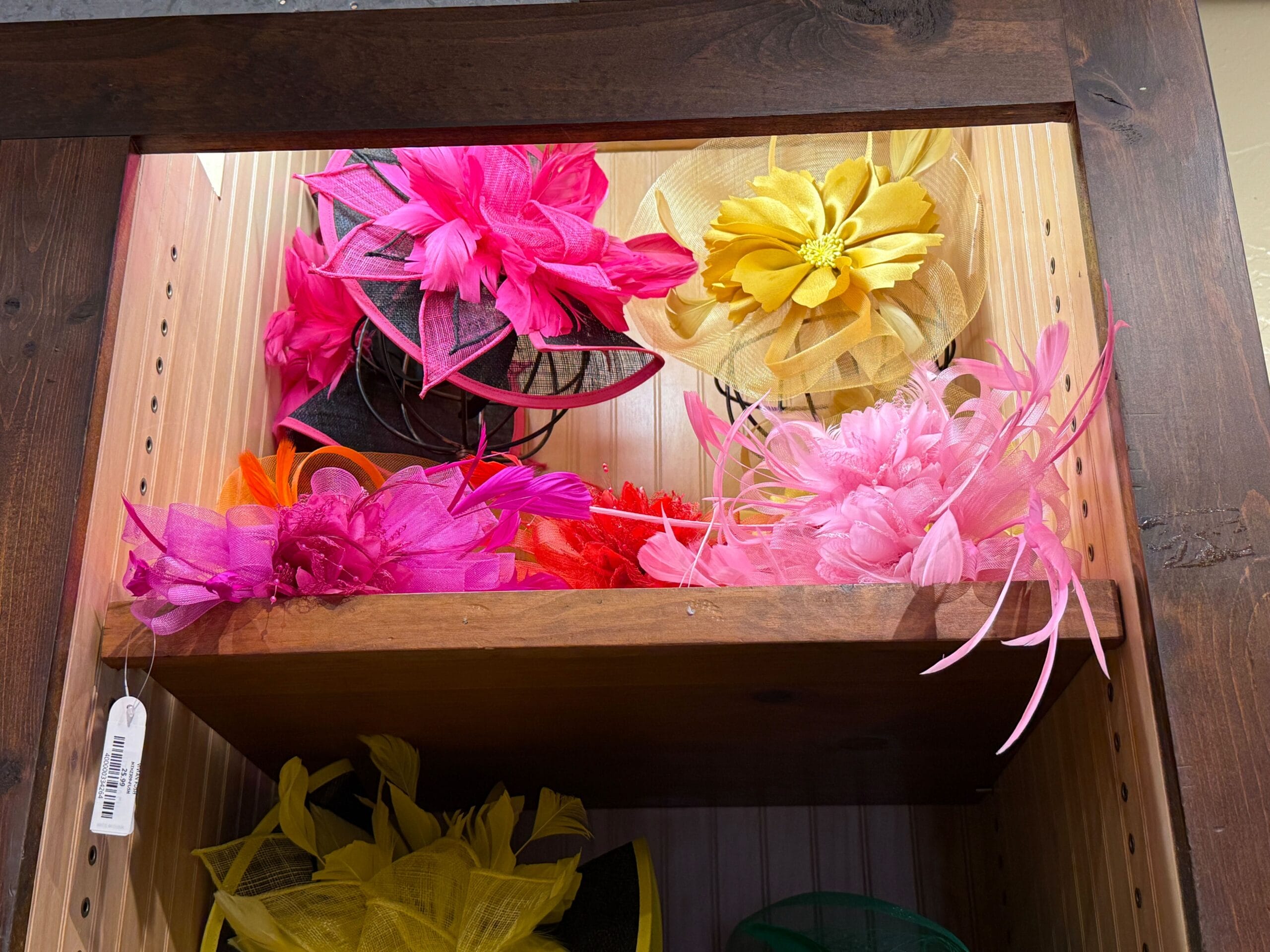 Brightly colored fascinators in pink, yellow, and orange line the shelves of the Prairie Outpost hat shop cabinet.