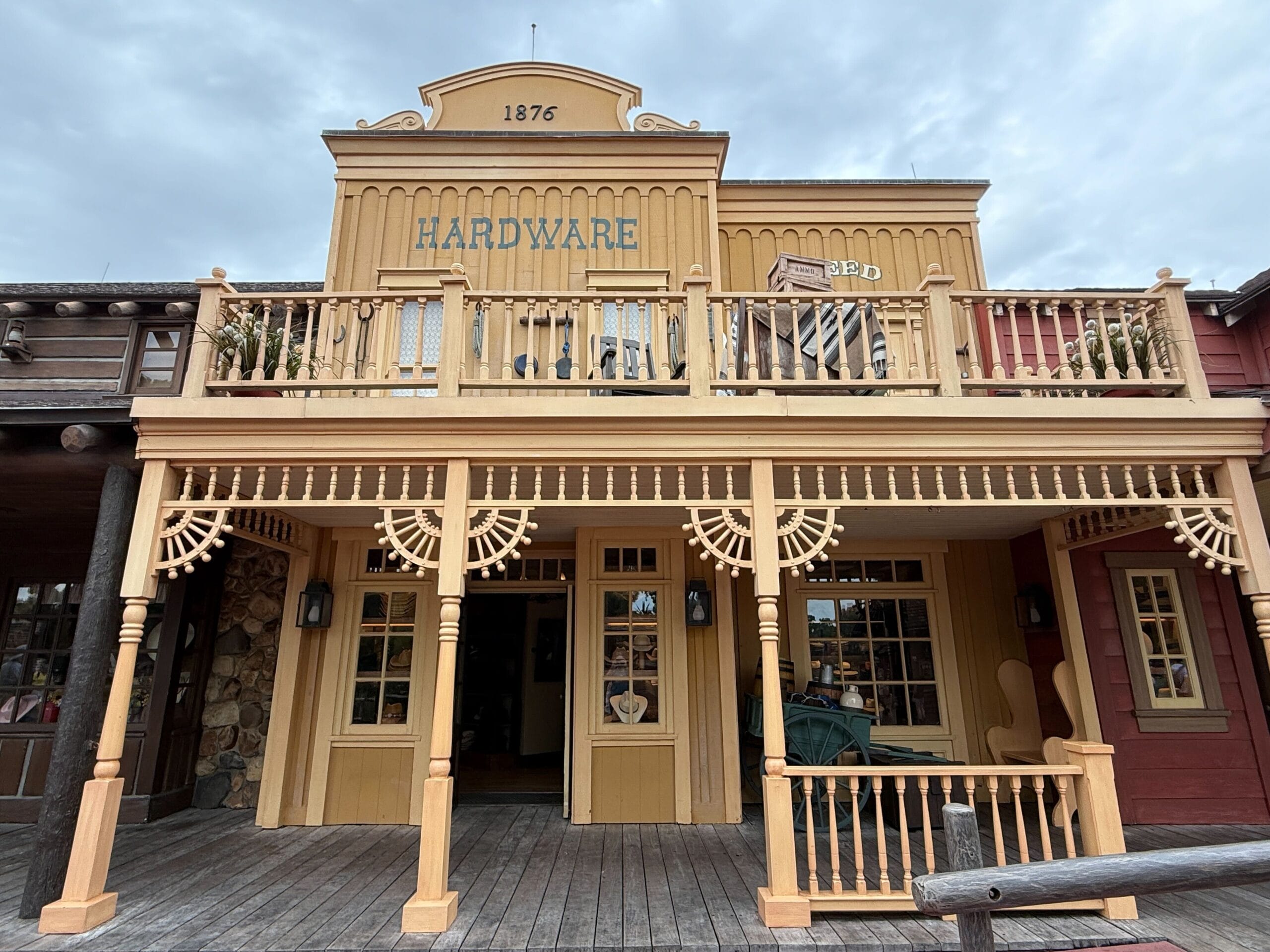 A yellow wooden Prairie Outpost hardware store with a balcony, labeled "1876 HARDWARE," under a cloudy sky.