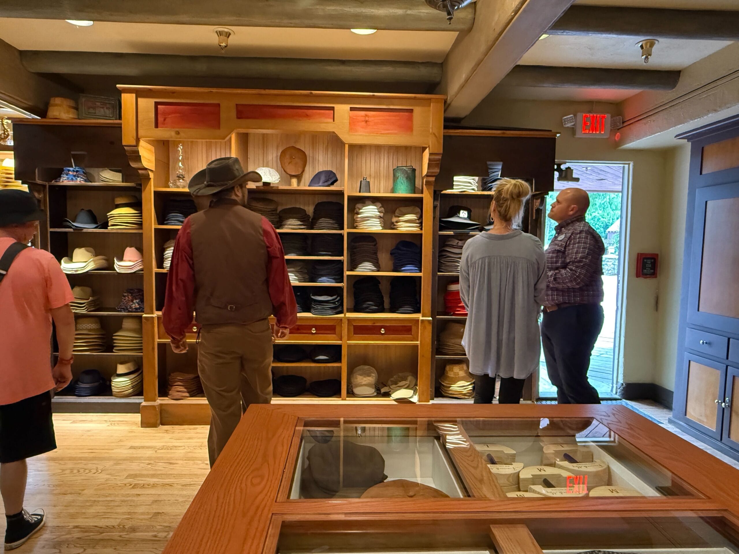 Four people browse the Prairie Outpost Hat Shop, admiring colorful hats on wooden shelves in Magic Kingdom.