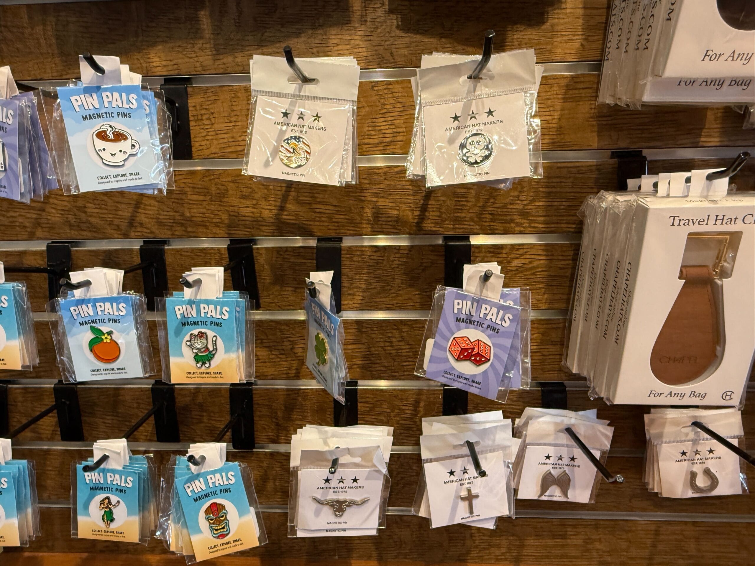 Colorful enamel pins and travel hat clips on display at the Prairie Outpost Hat Shop’s wooden wall rack.