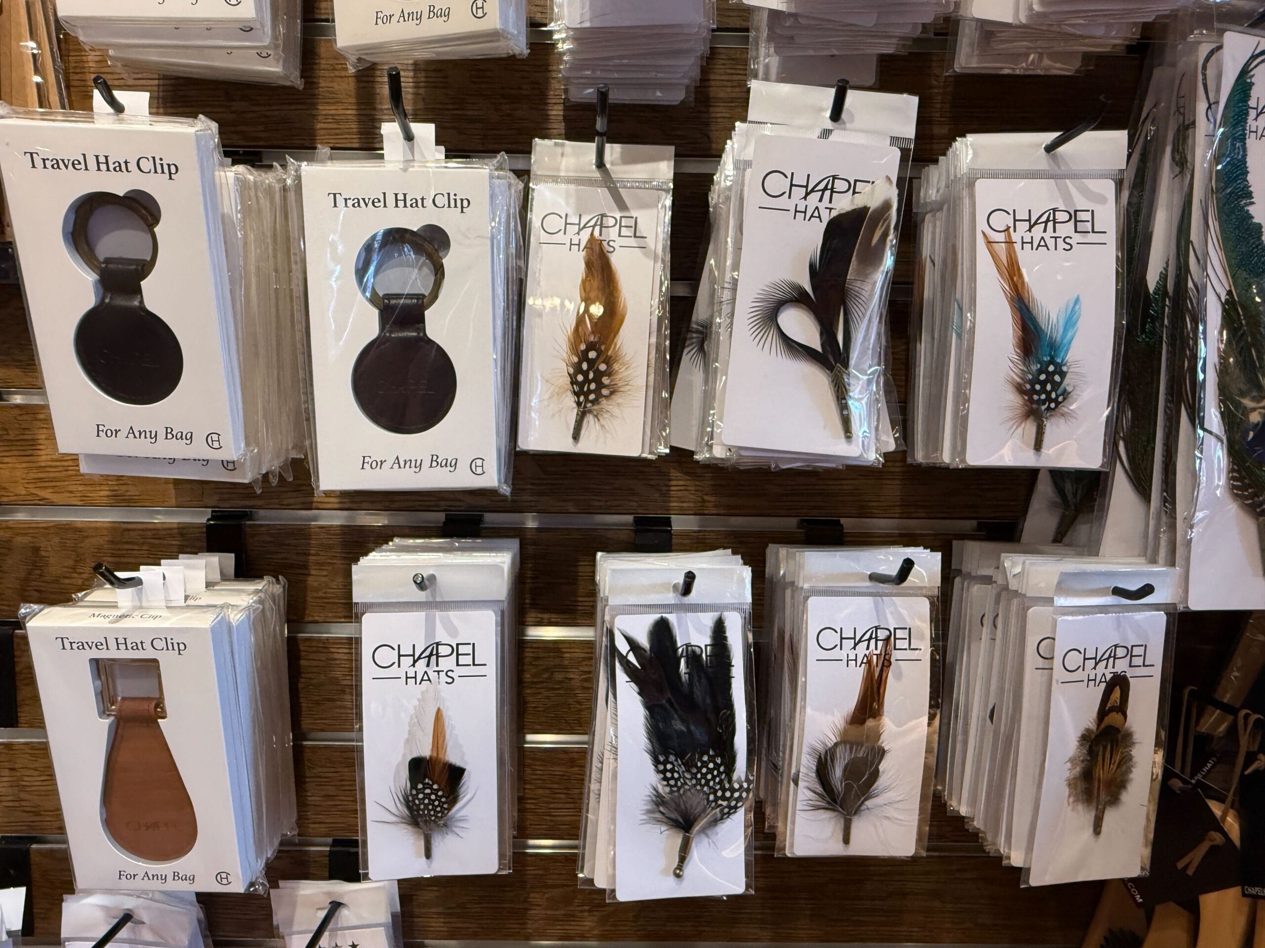 Packets of travel hat clips and feather hat accessories at the Prairie Outpost Hat Shop, displayed on a wooden wall rack.