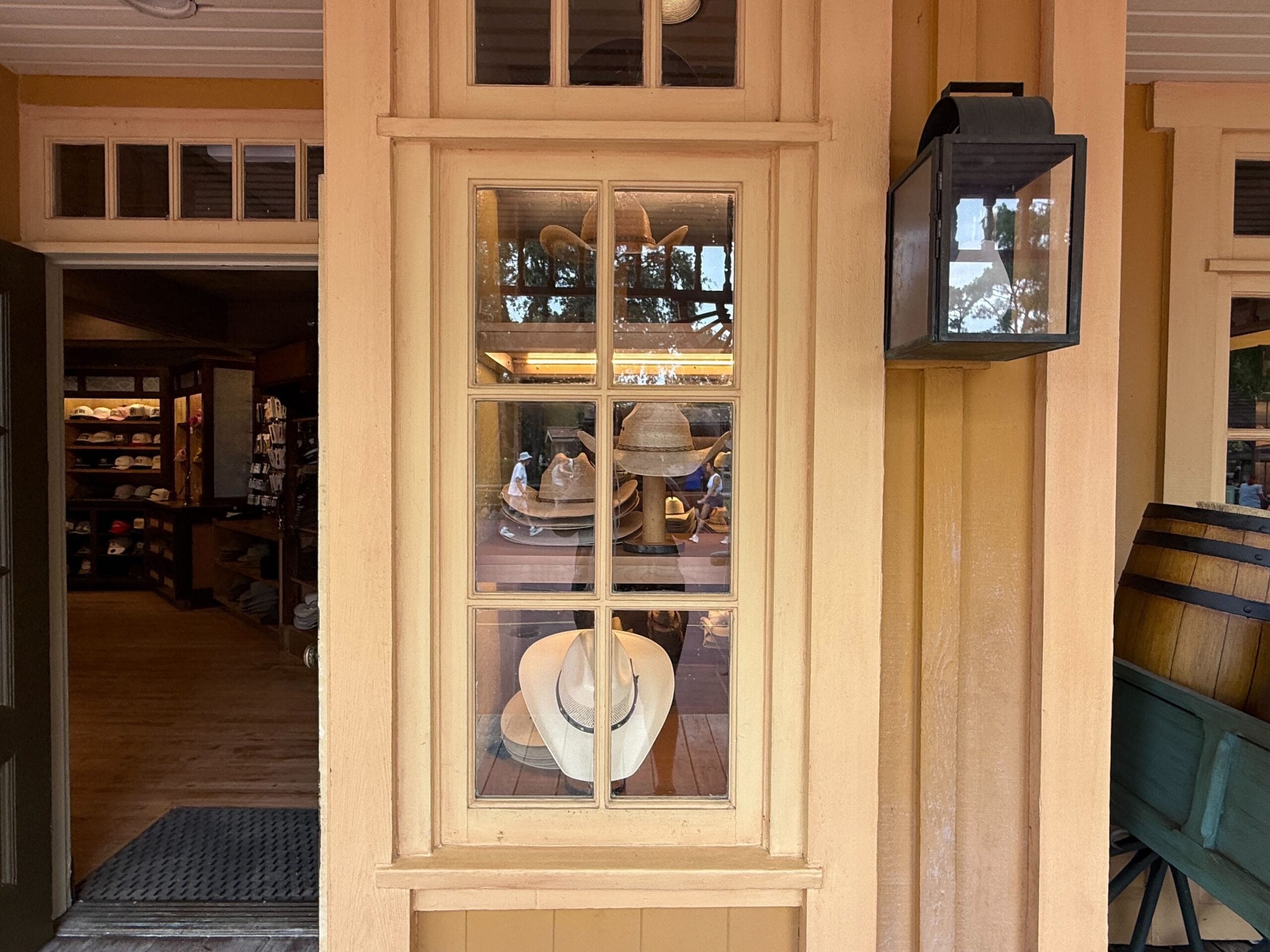 Display window with hats at Prairie Outpost, reflecting trees, beside a lantern and barrel outside a wooden Hat Shop.