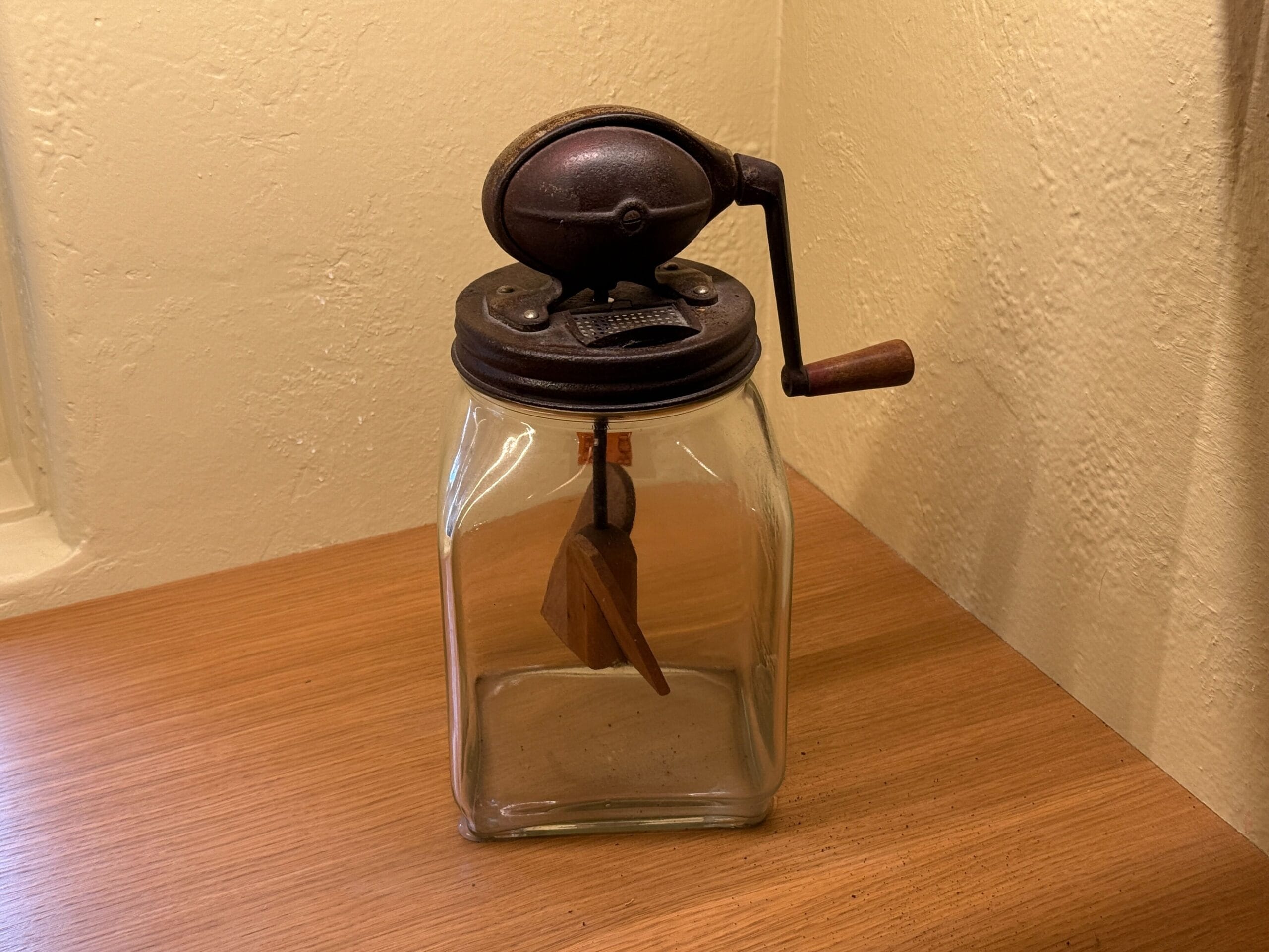 A vintage glass butter churn from Prairie Outpost rests on a wooden surface against a cream-colored wall.